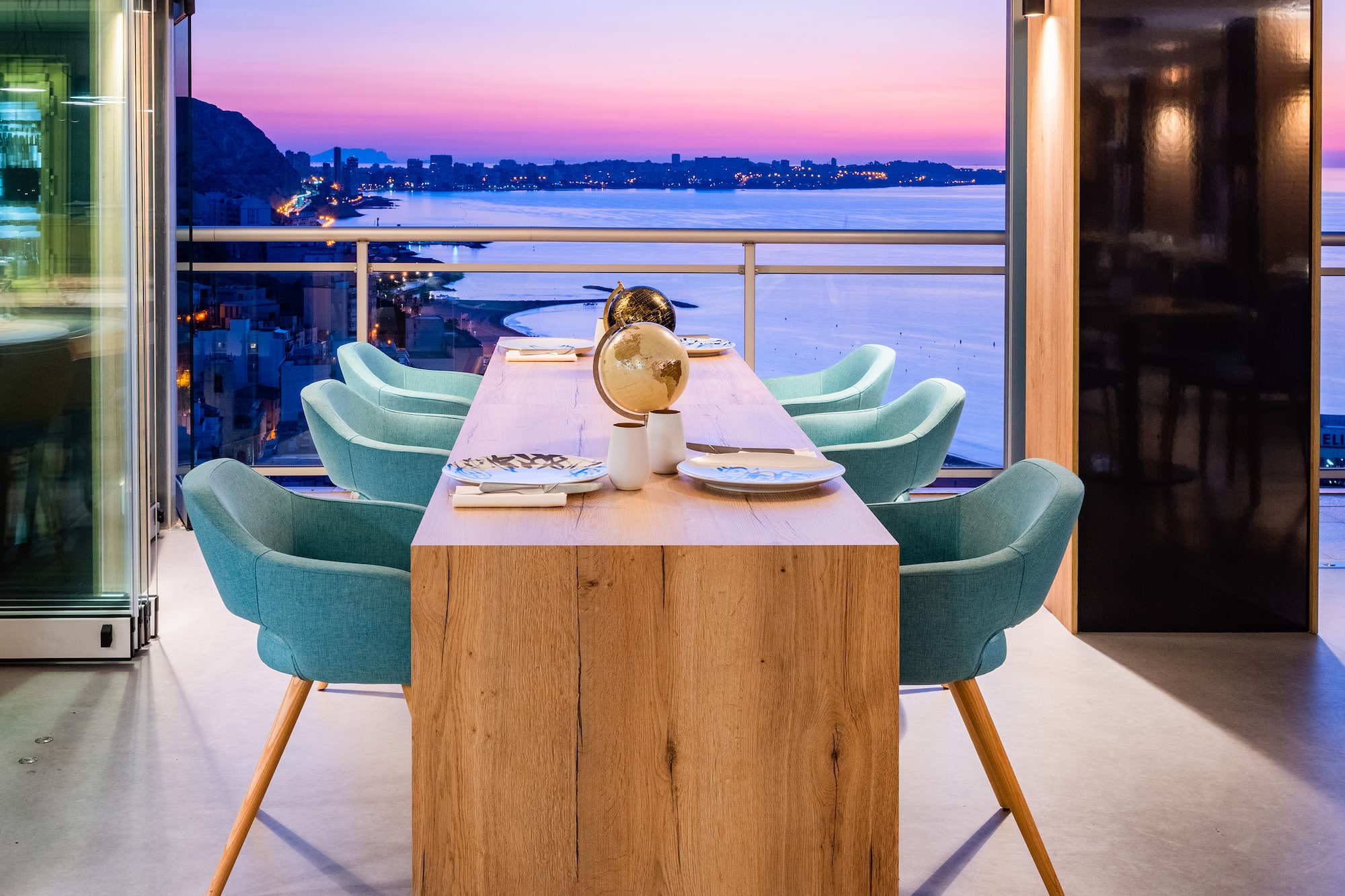 Modern dining table with teal chairs overlooking a coastal city at sunset.
