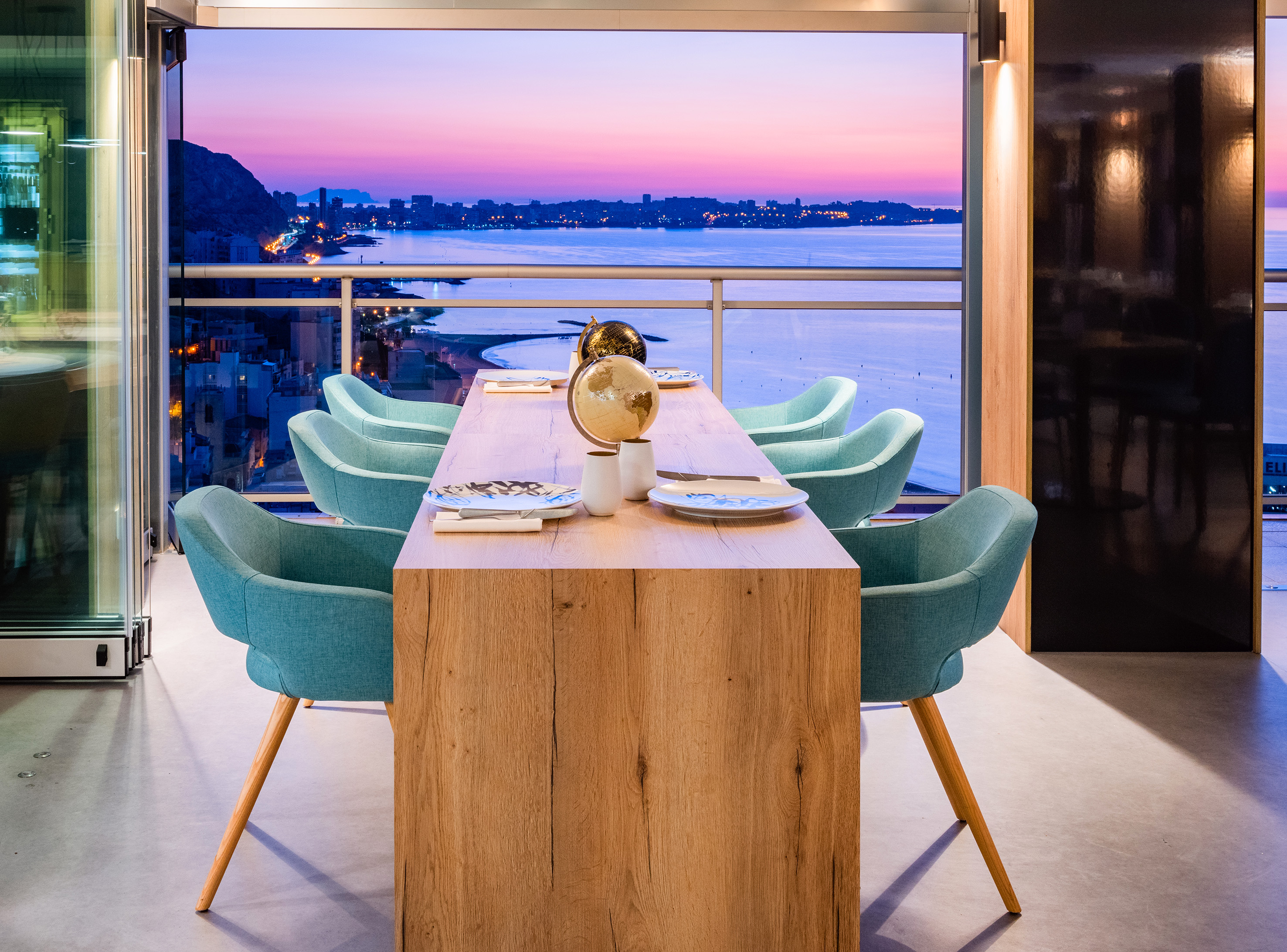 Modern dining table with teal chairs overlooking a coastal city at sunset.