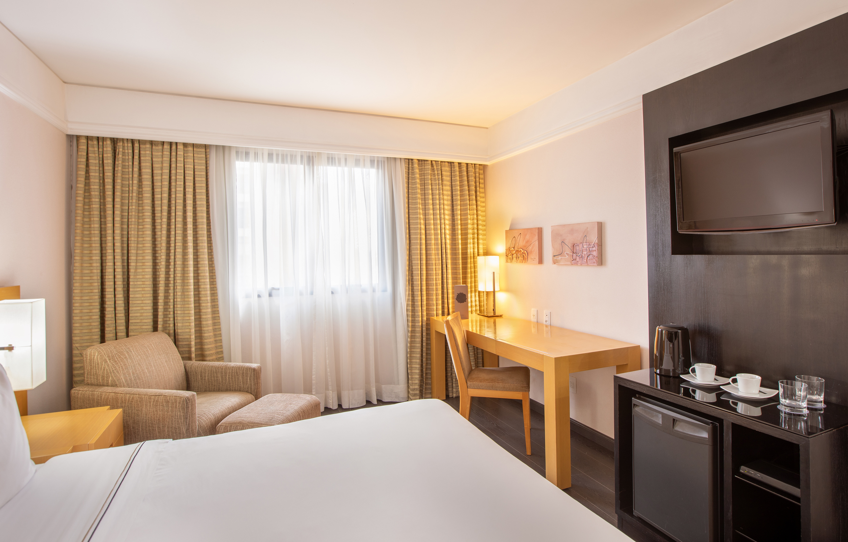 Hotel guest room with bed, desk, TV, mini-fridge, and armchair.
