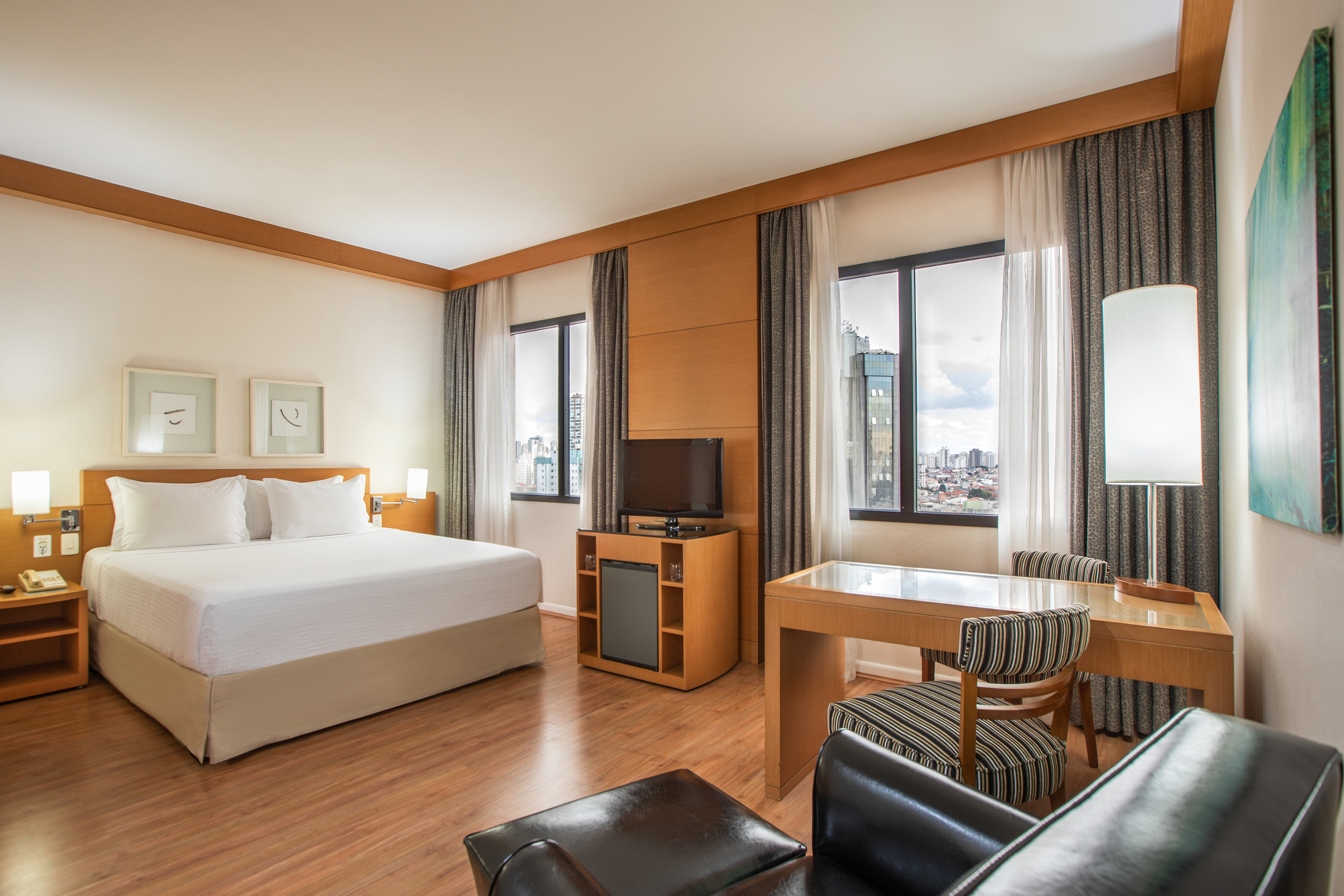 Modern hotel guest room with king bed, desk, TV, and city view windows.