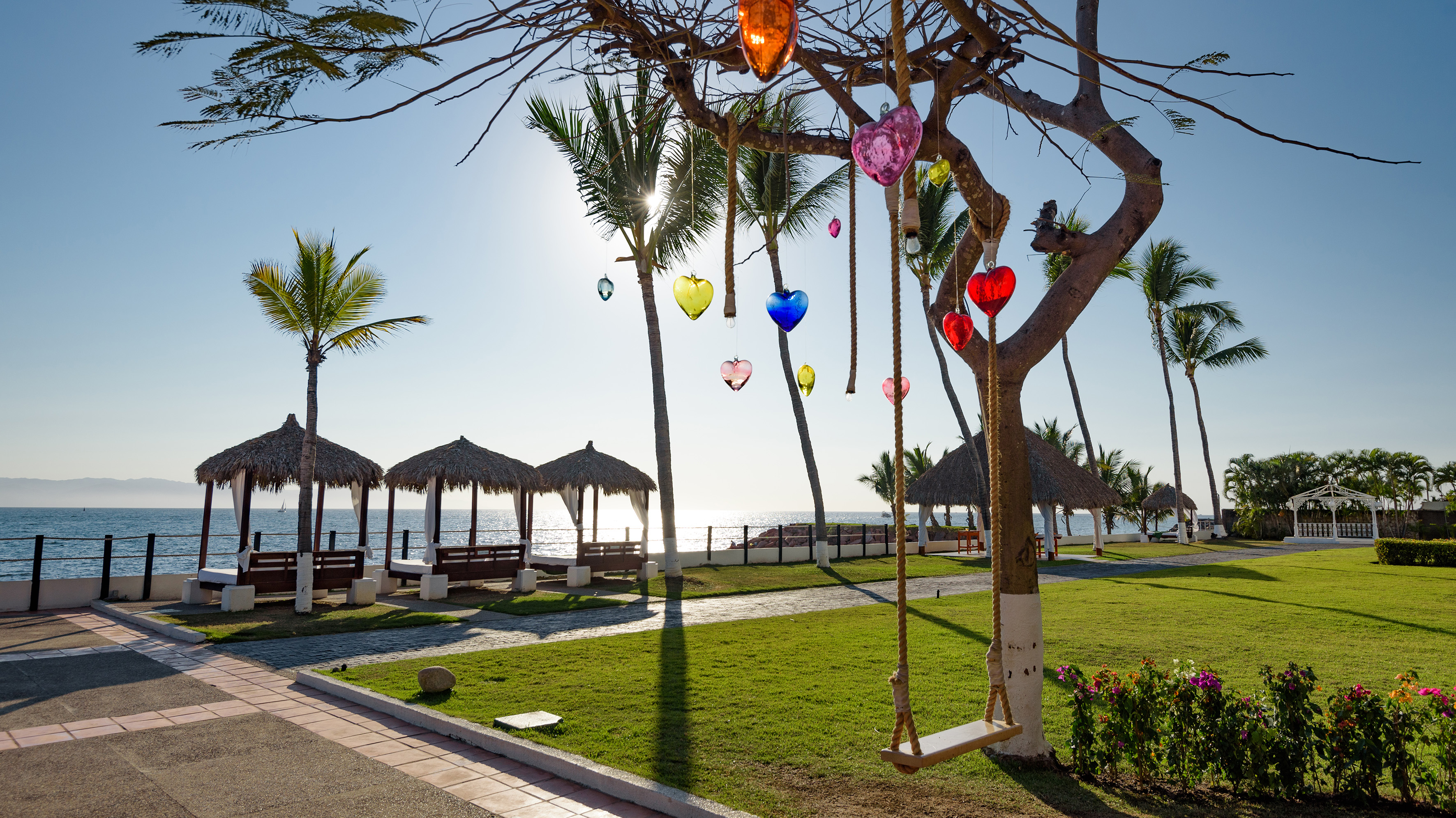 Tropical resort beach: heart ornaments, swing, palm trees, ocean, thatch cabanas.