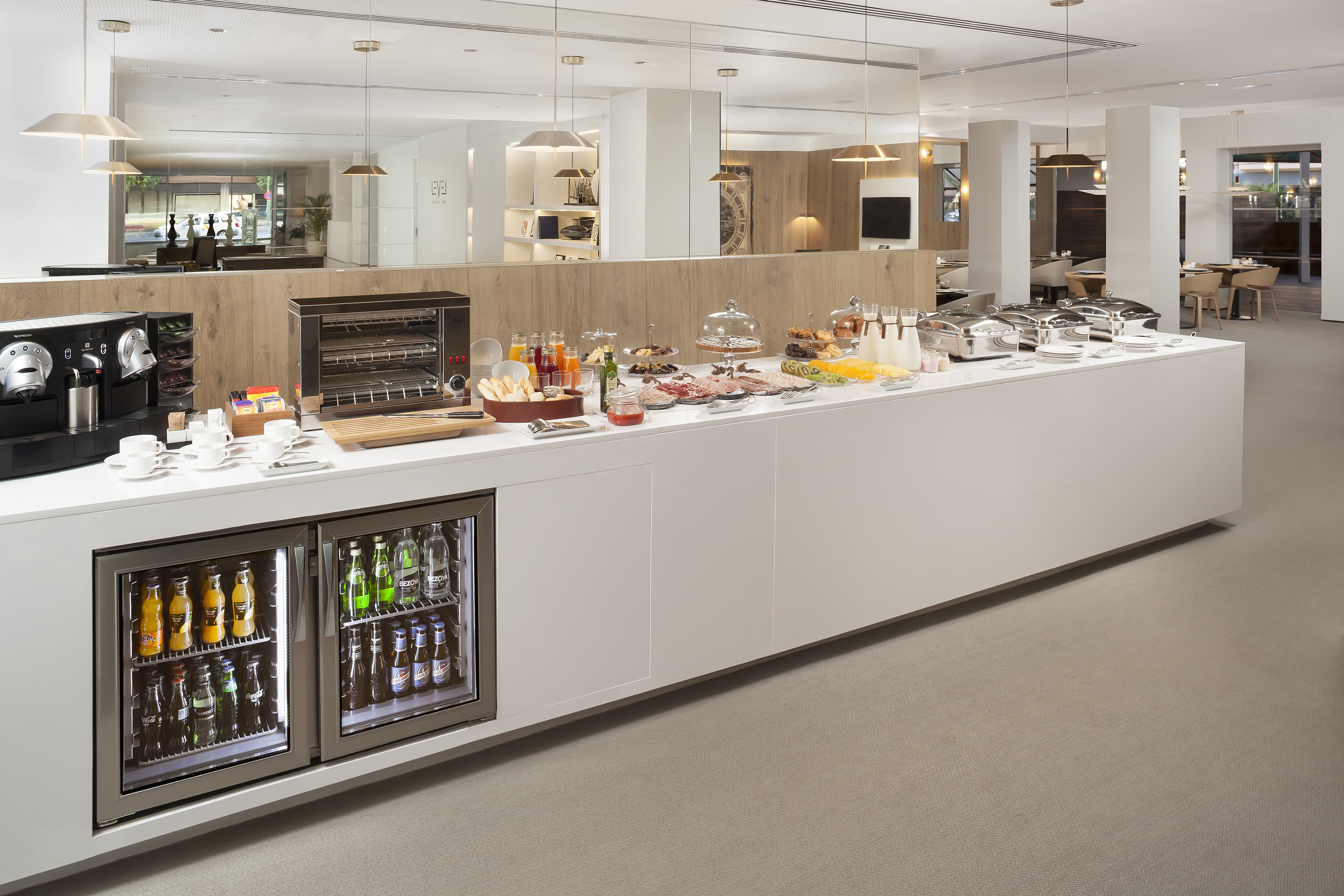 Modern hotel breakfast buffet with coffee, cold cuts, toaster, and varied drinks.