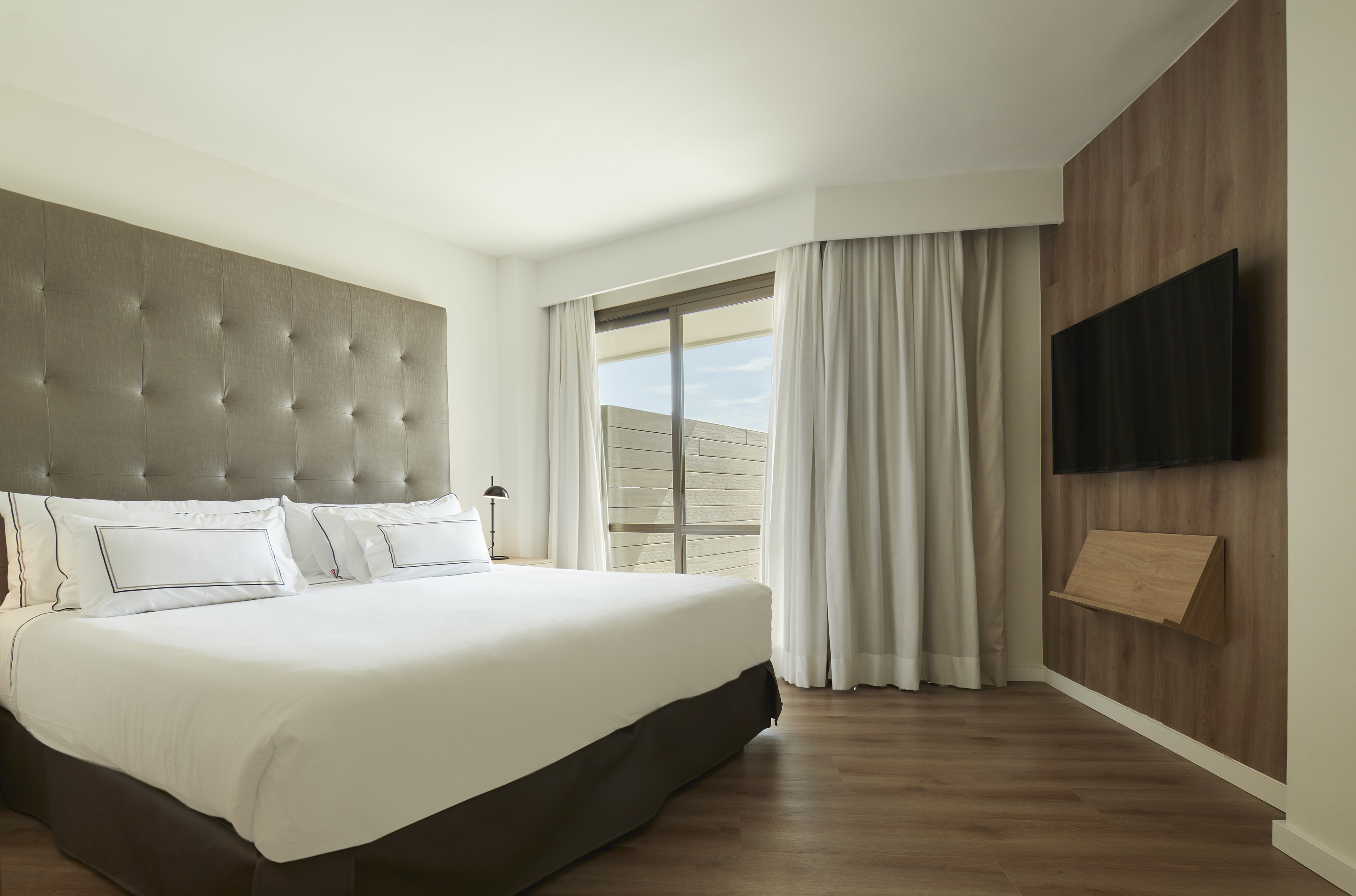 Modern hotel bedroom, king bed, tufted headboard, window, and wall TV.