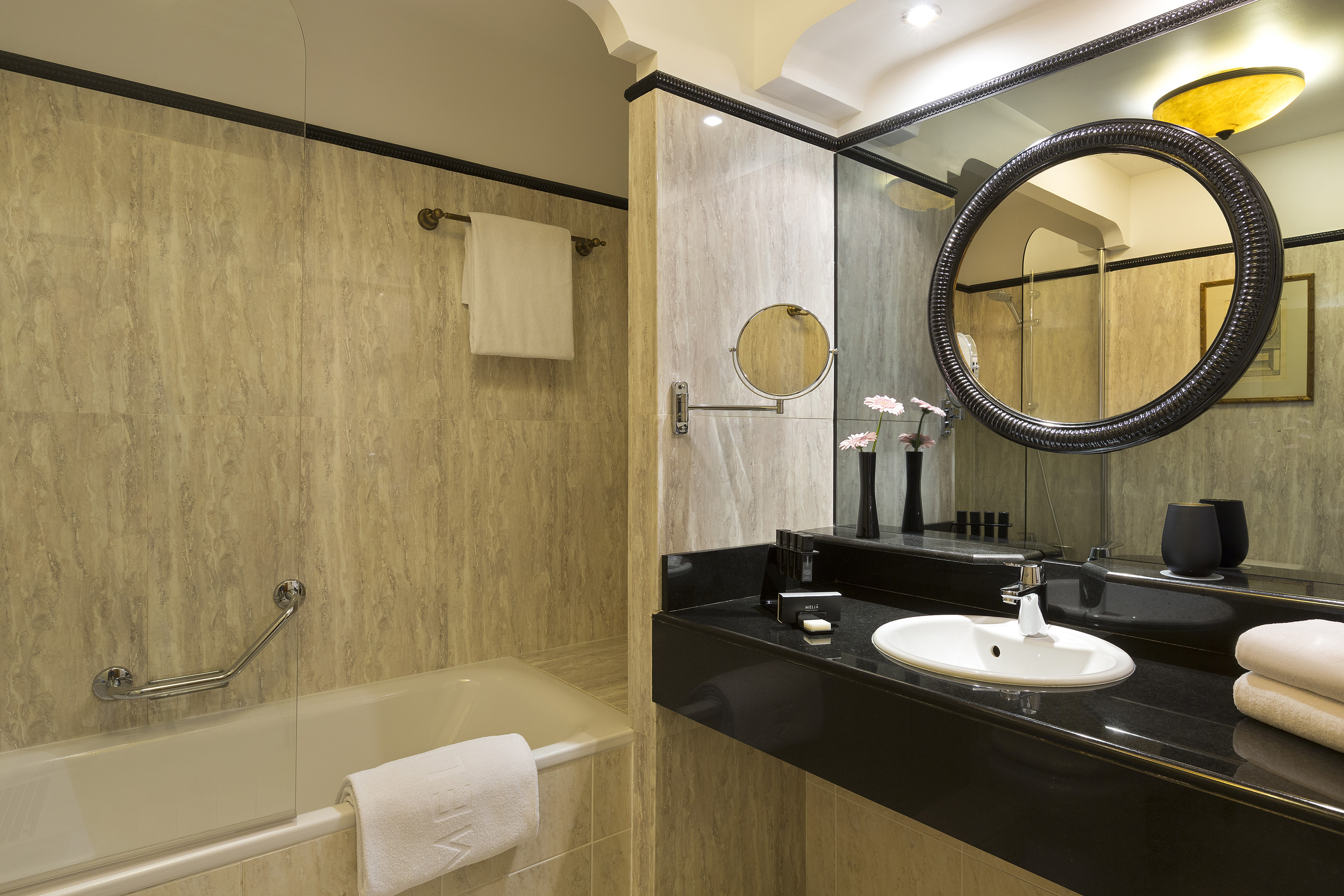 Hotel bathroom with black vanity, round mirror, white sink, and tiled bathtub.