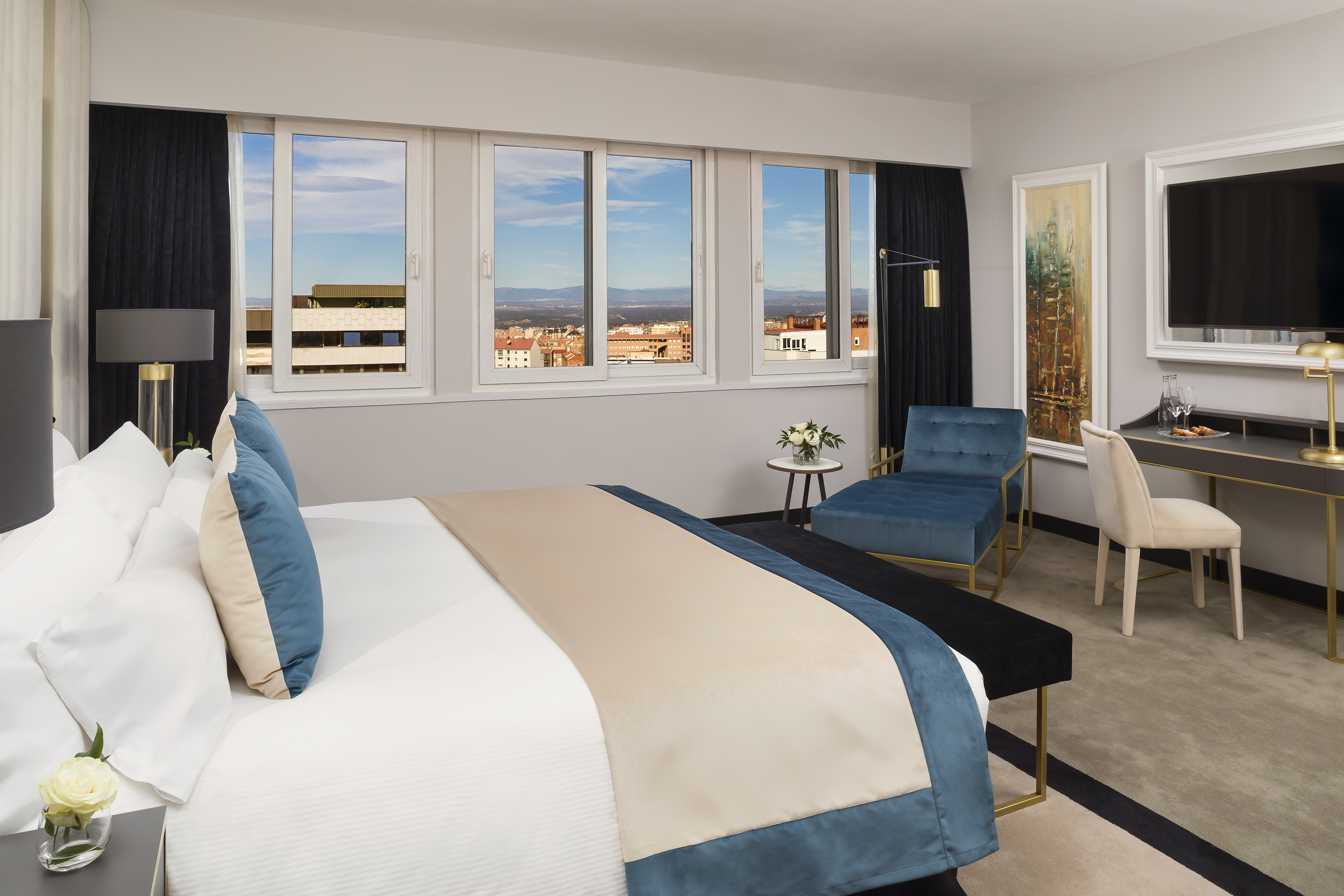 Modern hotel bedroom with king bed, city view, desk, and blue chaise lounge.