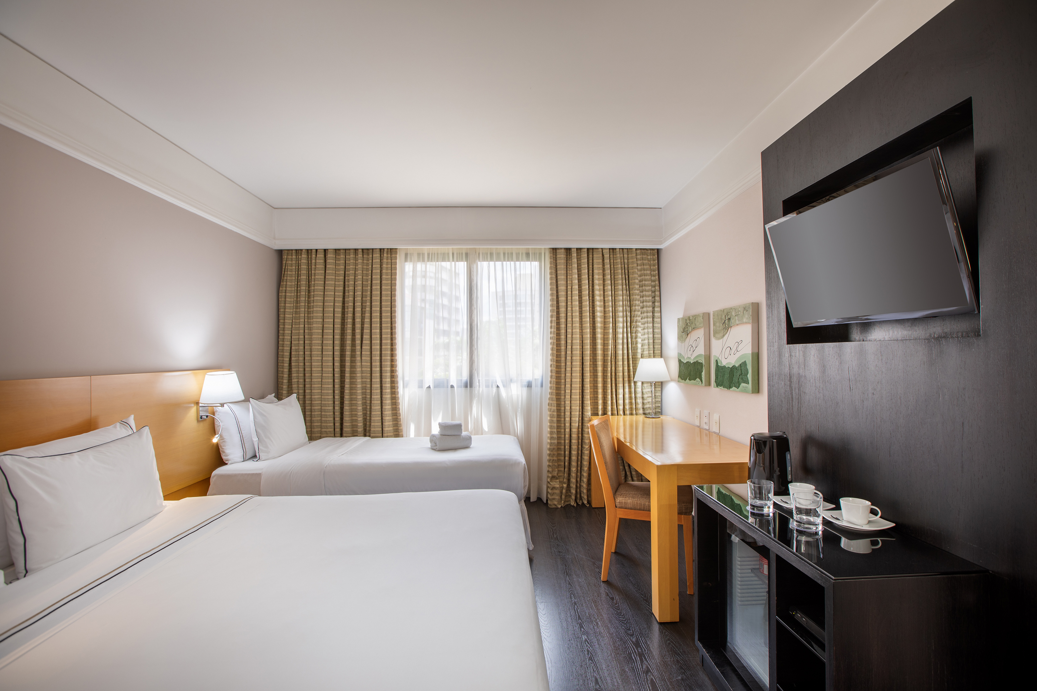 Hotel room with two twin beds, desk, wall-mounted TV, and sunlit window.