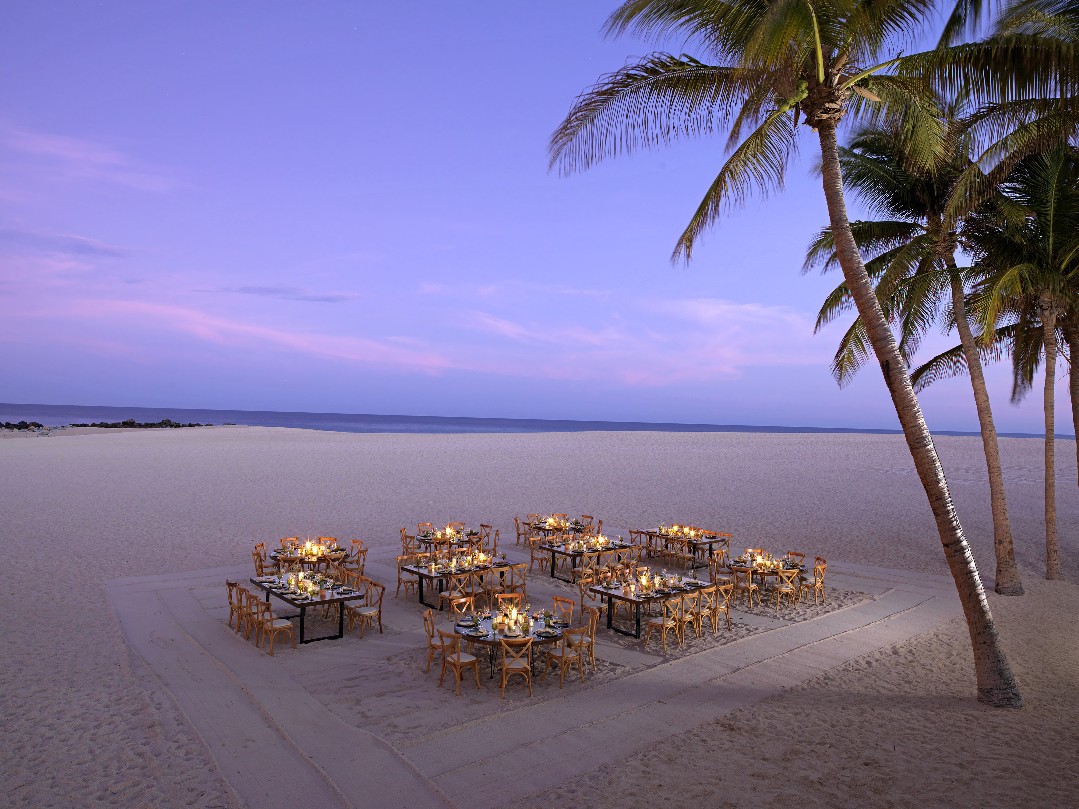 Luxury beach dining tables with candles, palm trees, and ocean at dusk.