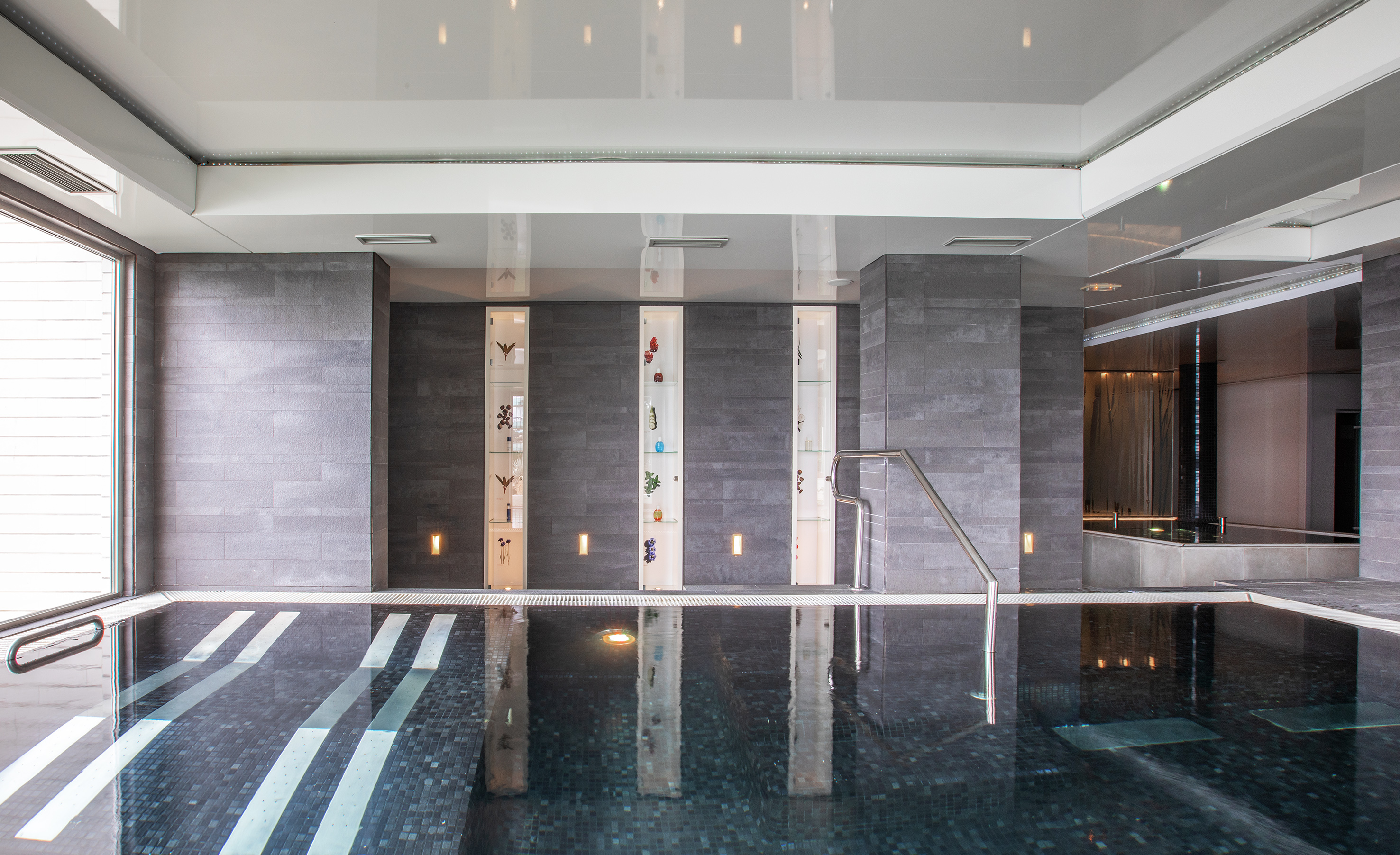 Modern indoor pool, dark mosaic tiles, grey walls, illuminated decorative shelves.