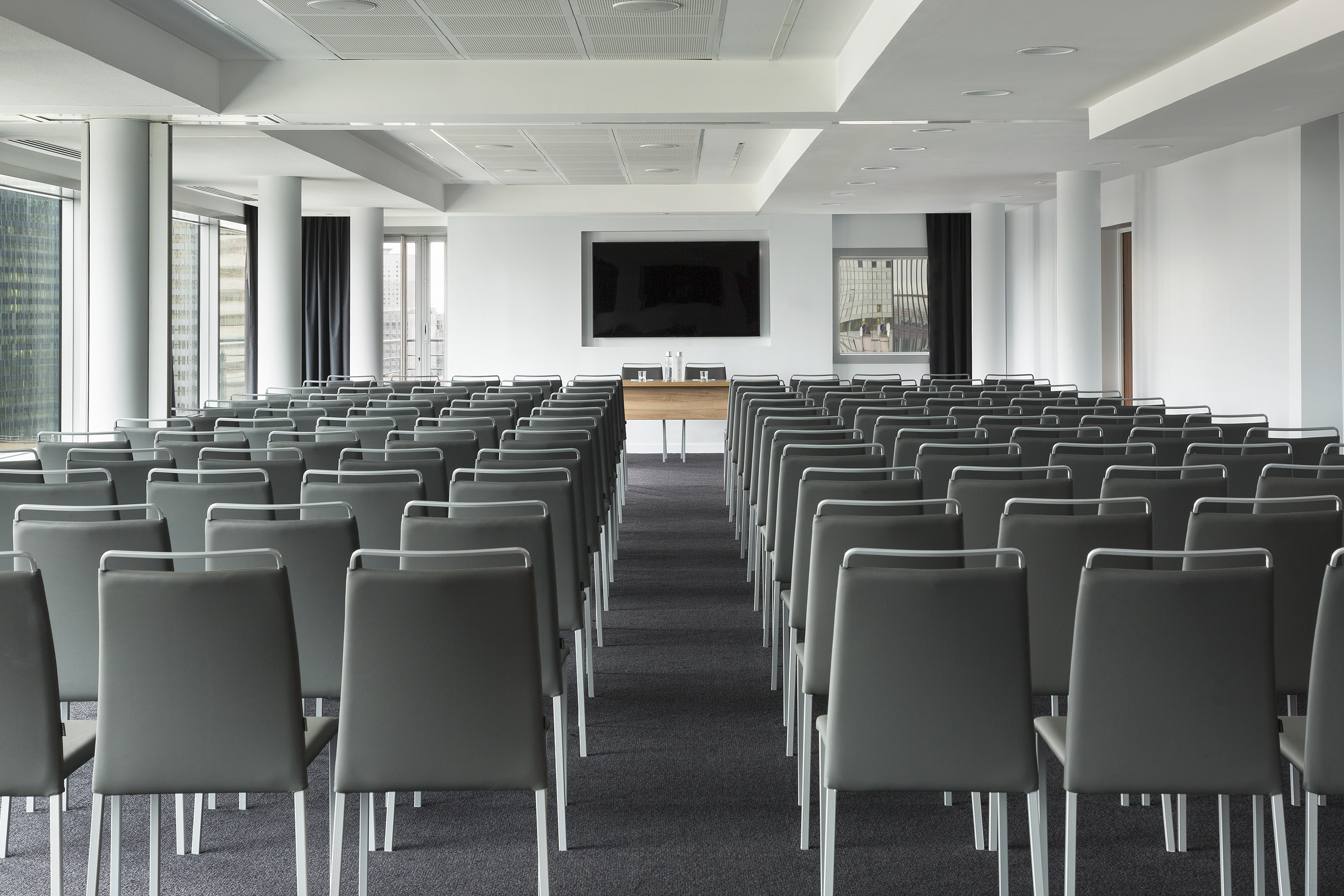 Empty modern conference room, rows of gray chairs facing large screen and table.