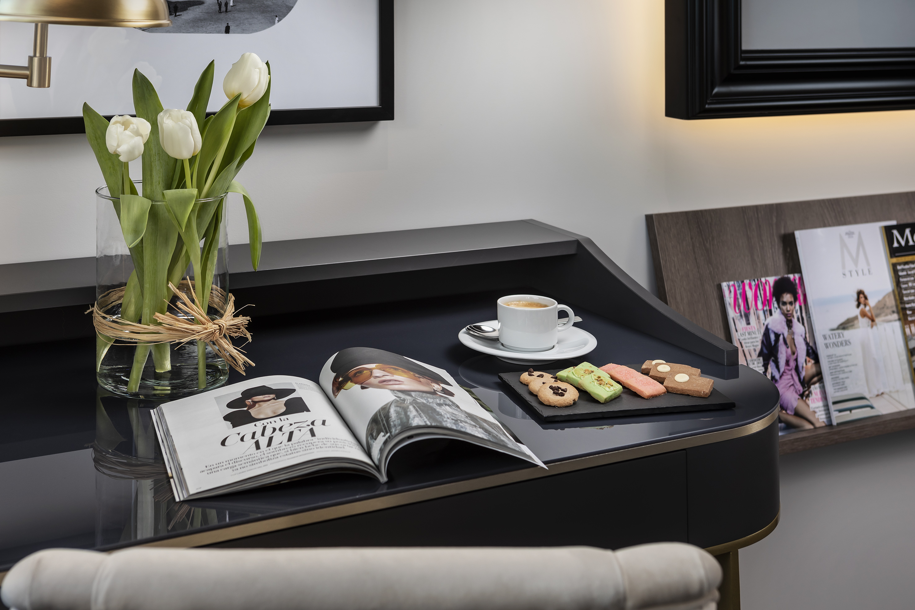 Elegant hotel room desk with white tulips, coffee, colorful cookies, and open magazine.