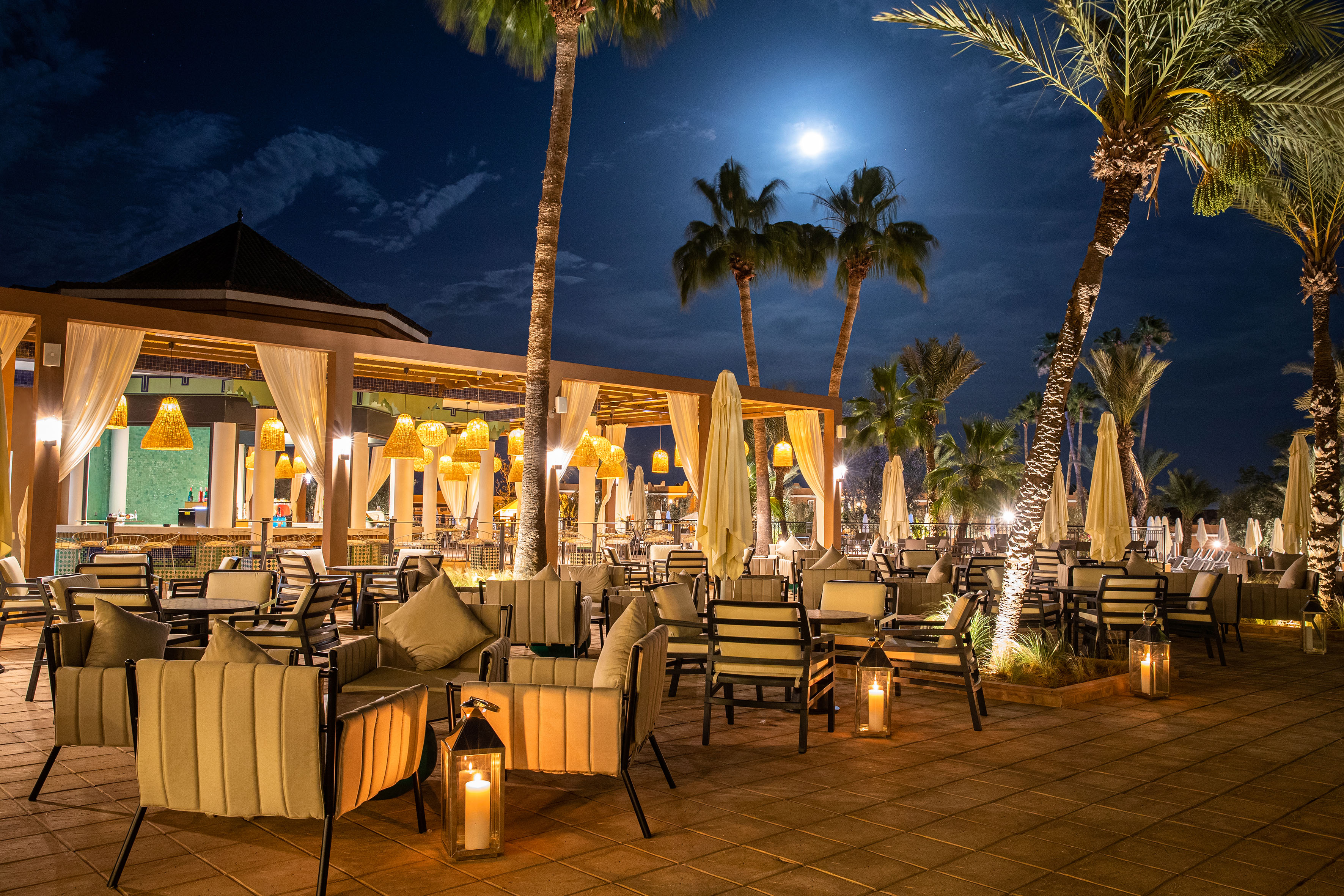 Nighttime resort patio: palm trees, full moon, warm lights, outdoor dining.
