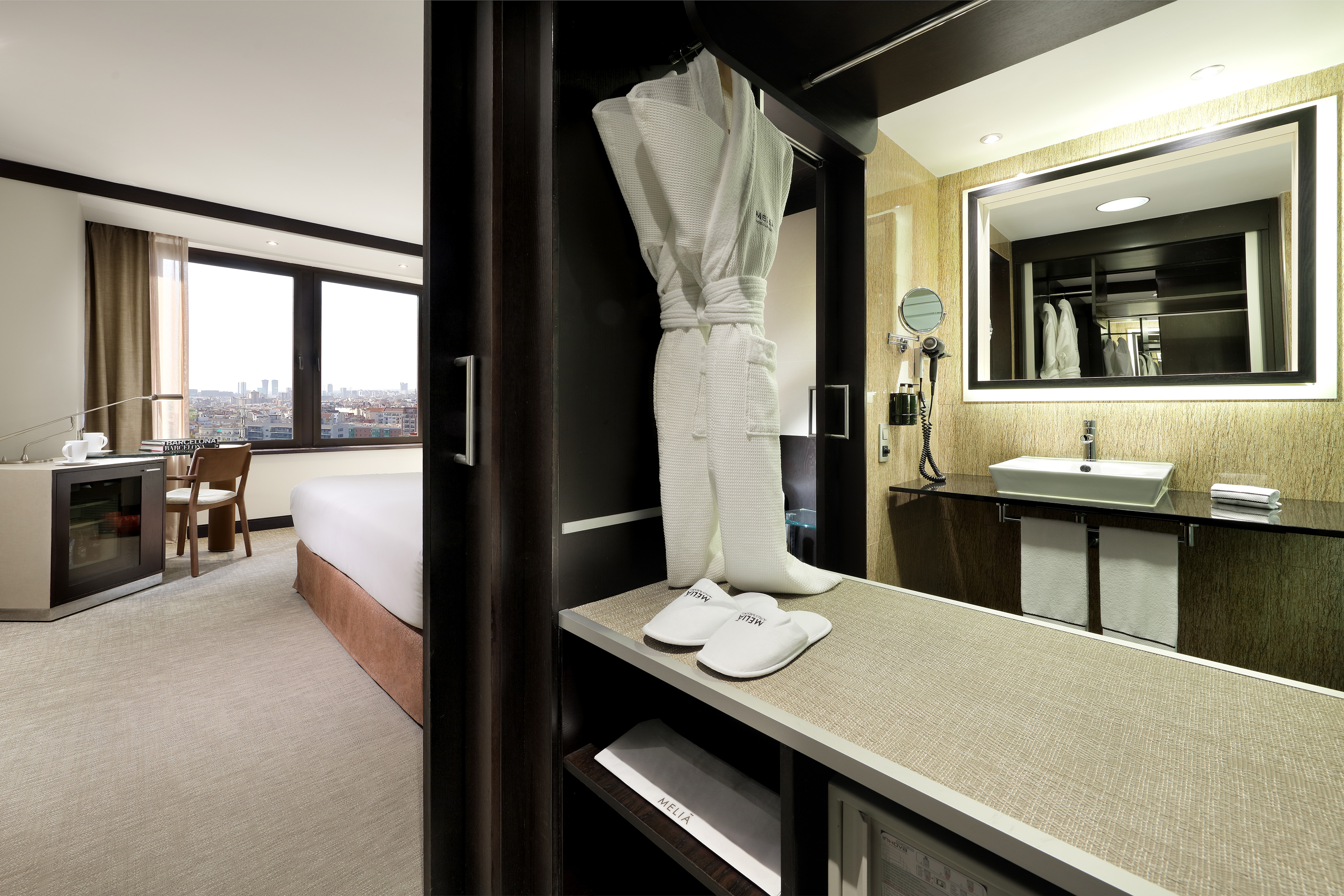 Hotel bedroom with city view, desk, and vanity with bathrobe and slippers.