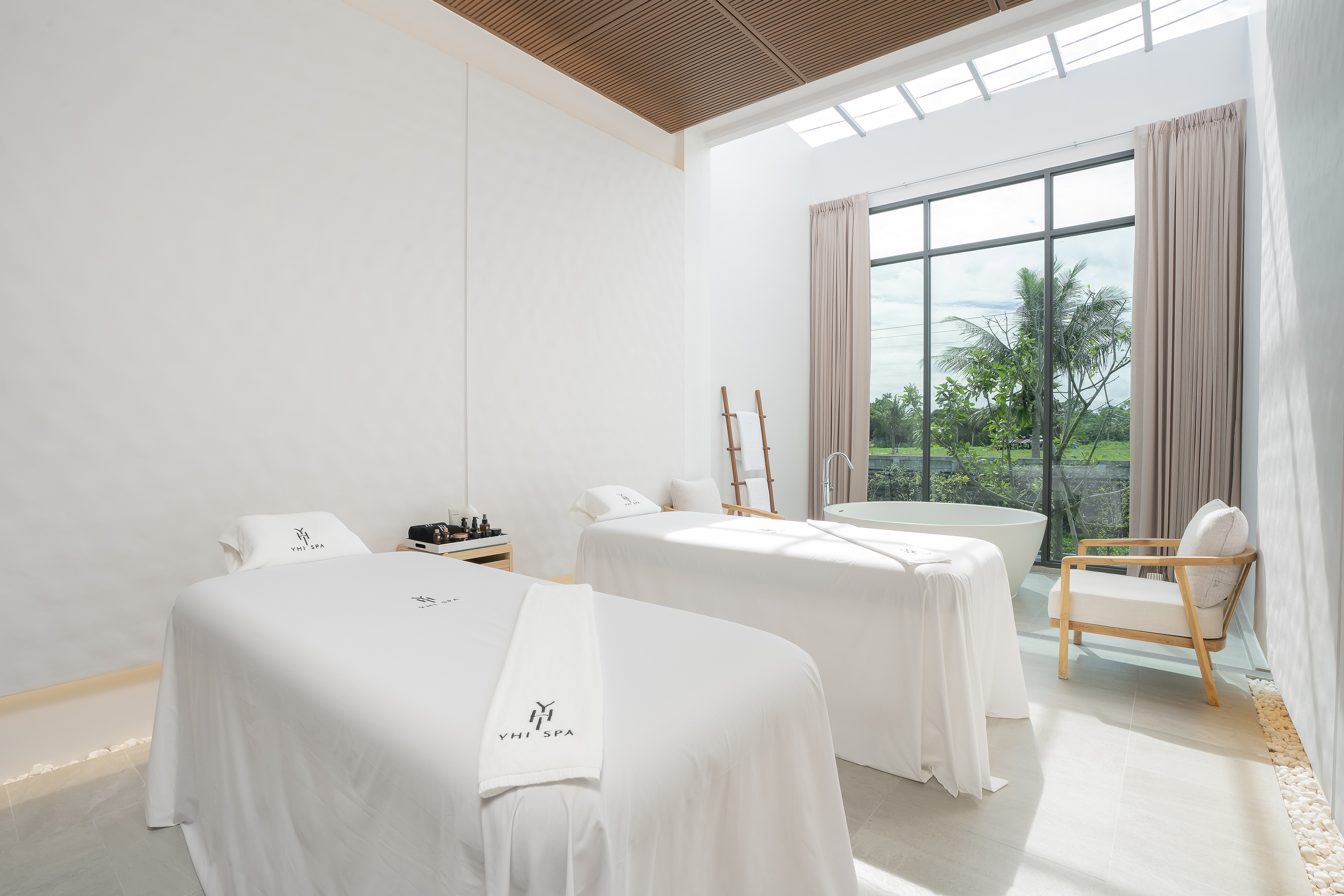 YHI Spa massage room, two beds, tropical window view, and bathtub.