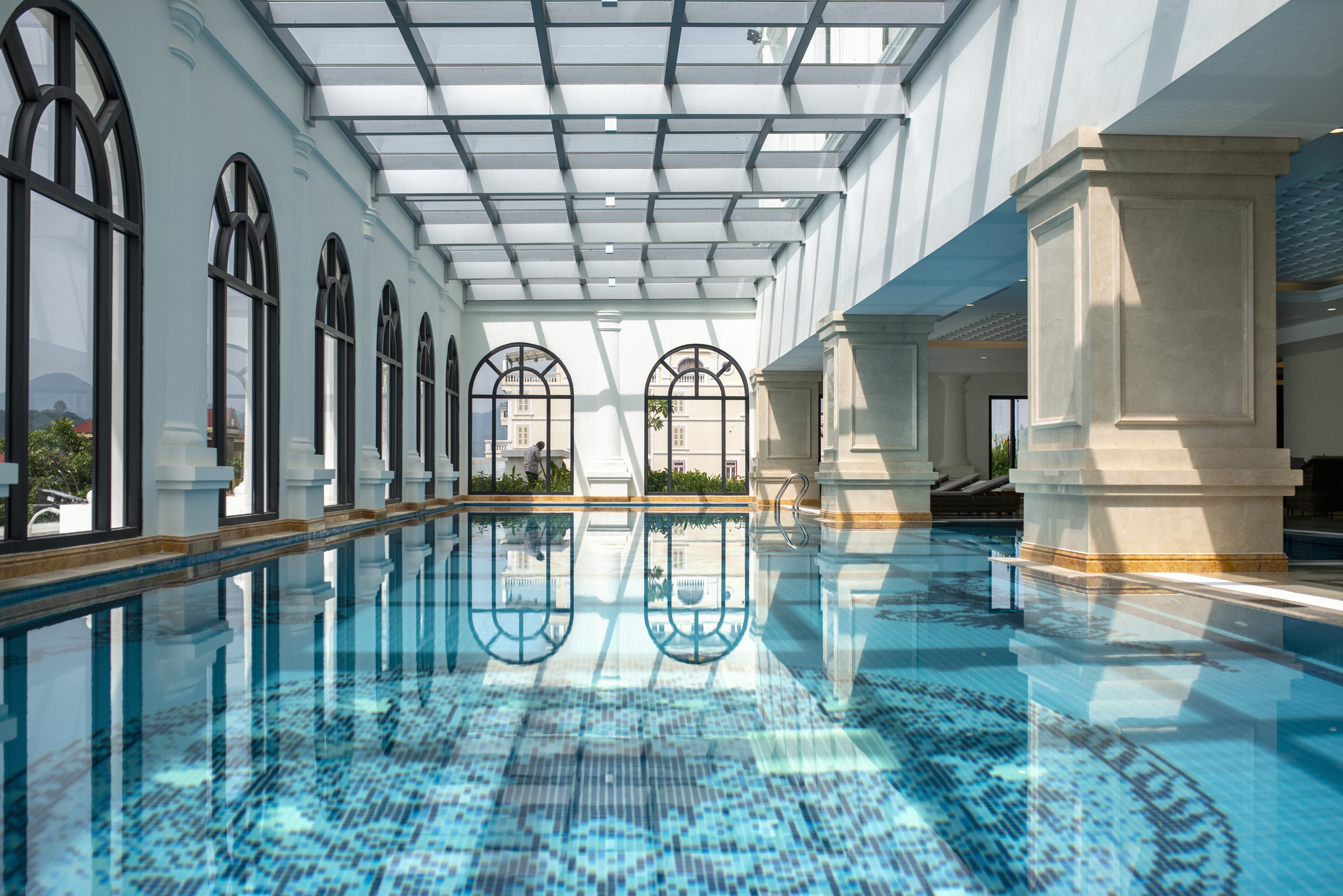 Indoor swimming pool with arched windows, glass ceiling, and blue mosaic tiles.