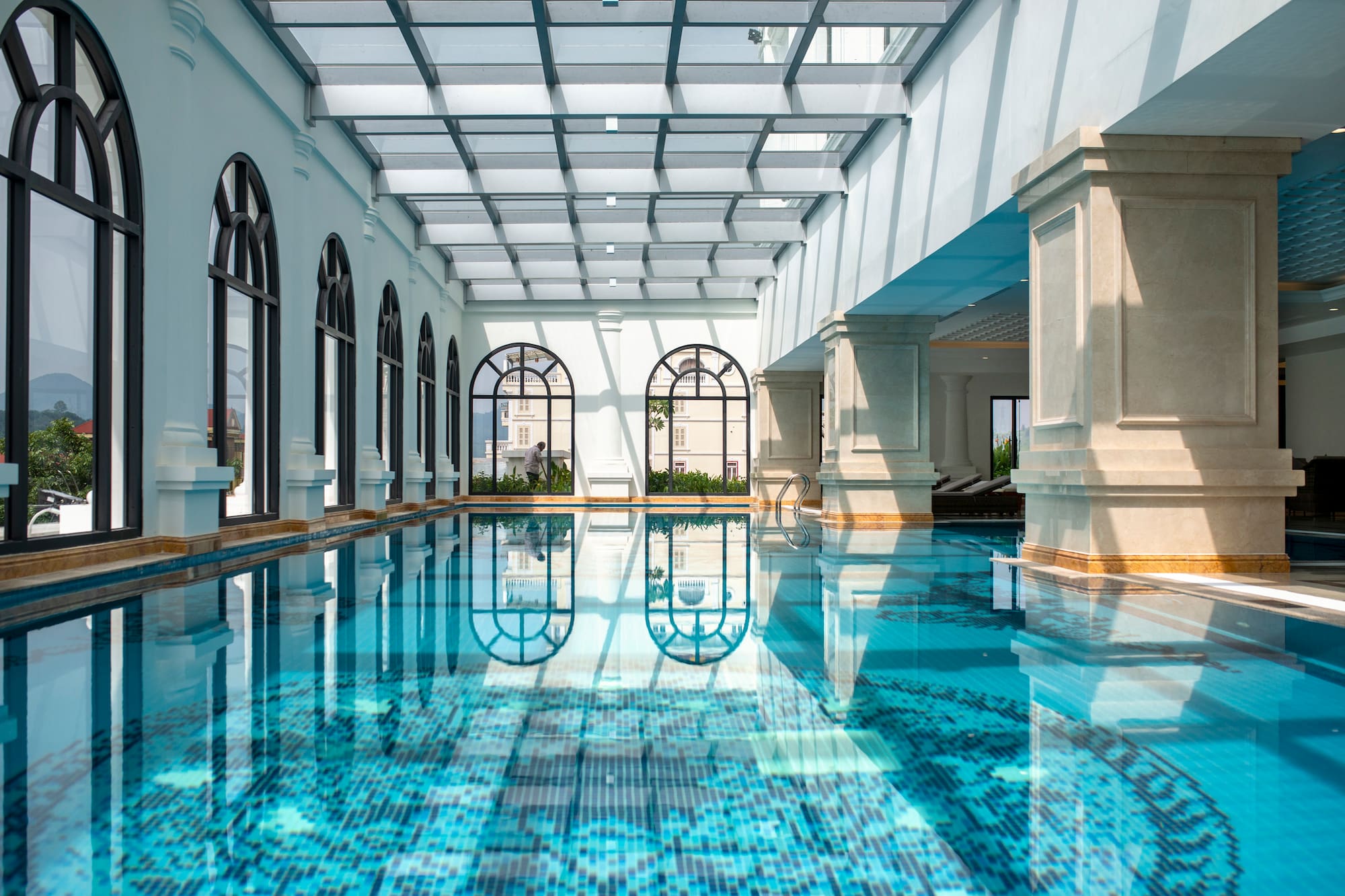 Indoor swimming pool with arched windows, glass ceiling, and blue mosaic tiles.