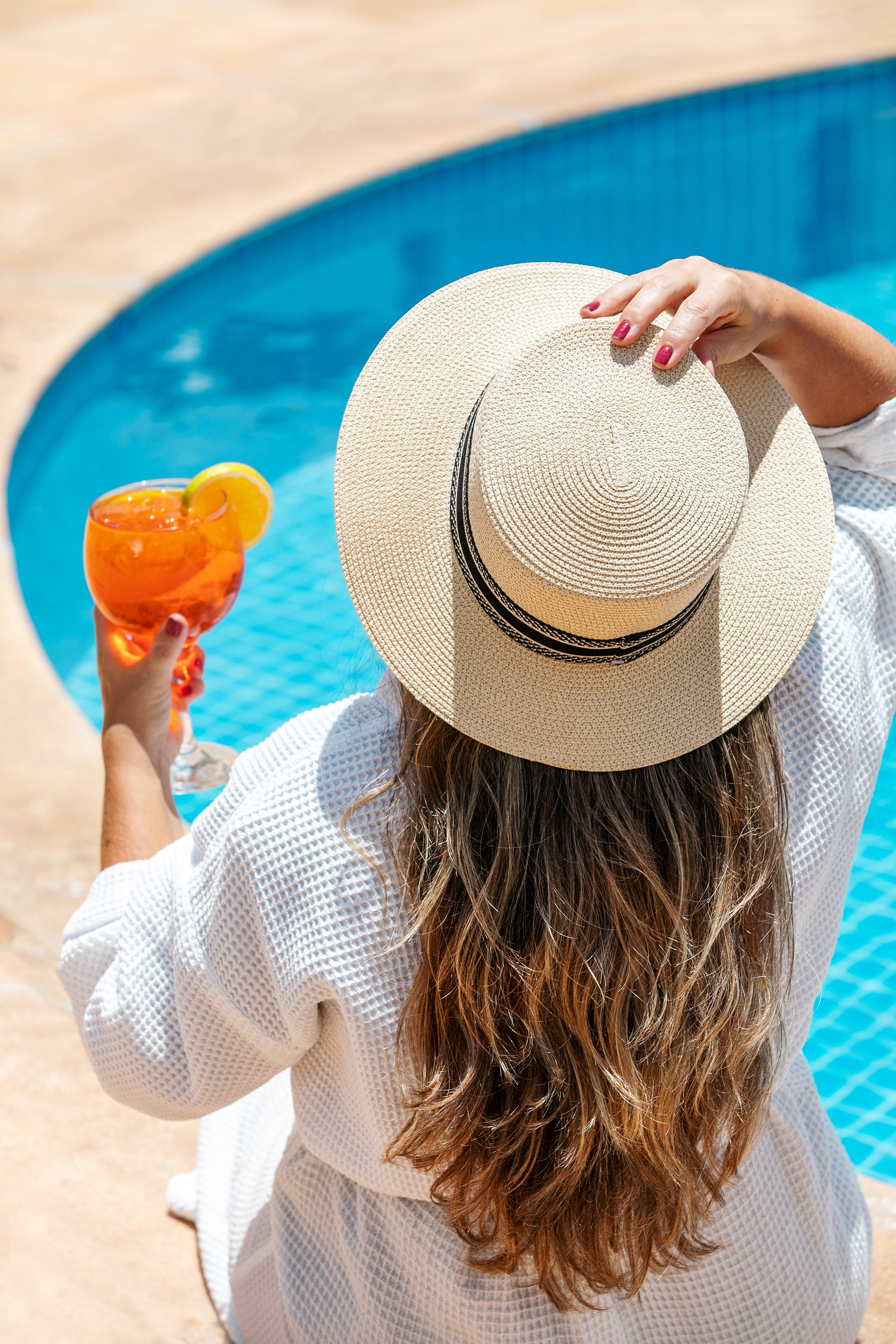 Back of woman in straw hat and robe, holding orange cocktail by sunny swimming pool.