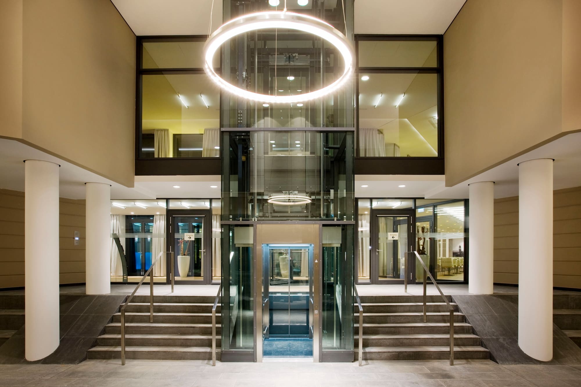 Modern lobby with glass elevator, circular light, stairs, and ramp access.