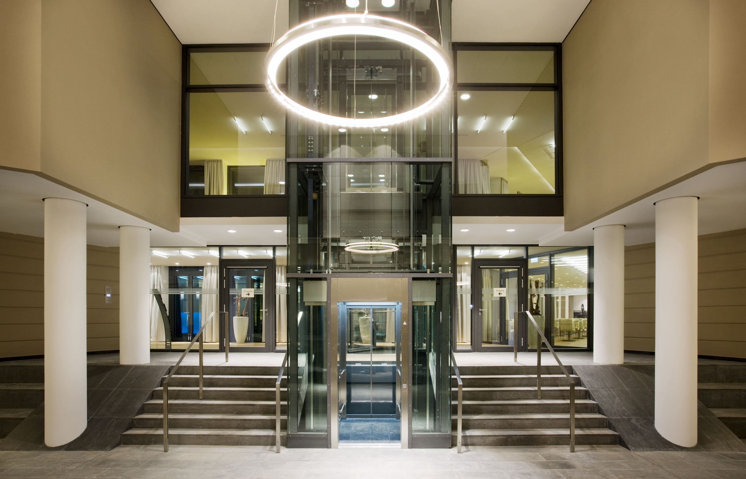 Modern lobby with glass elevator, circular light, stairs, and ramp access.