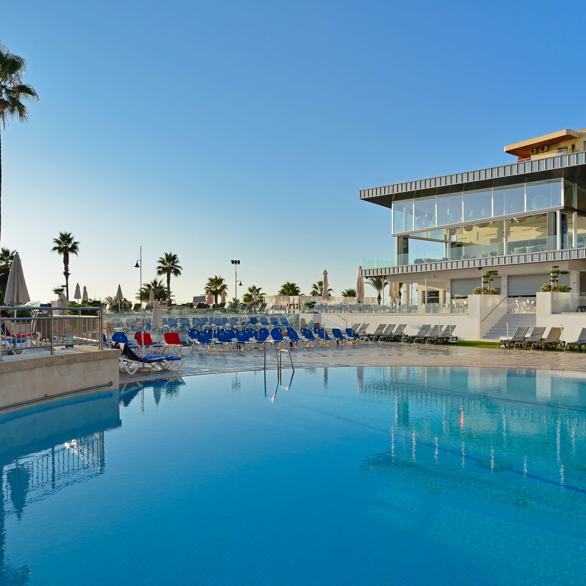 Modern hotel swimming pool with lounge chairs and palm trees under a blue sky.