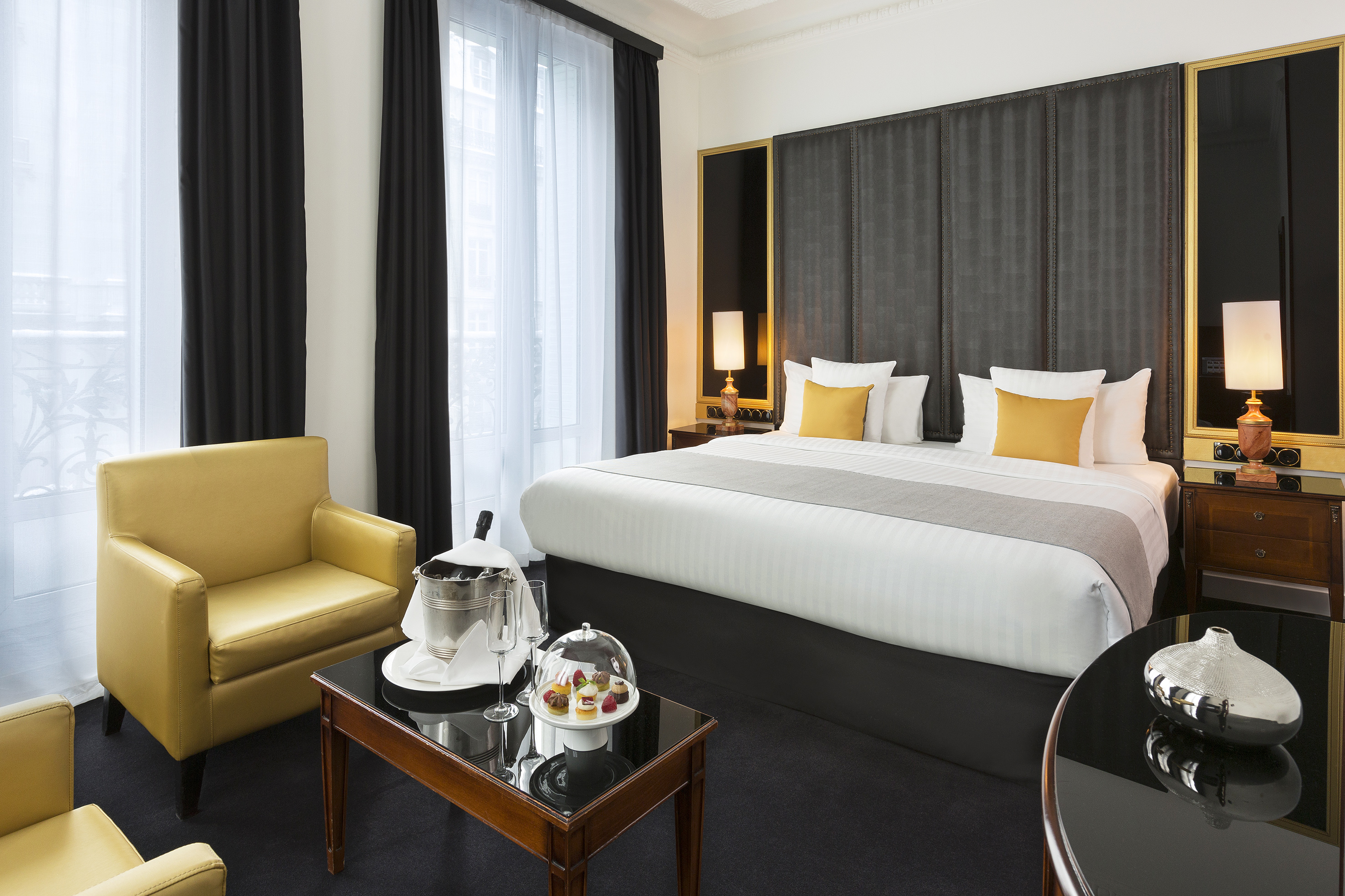 Elegant hotel room with king bed, yellow chair, champagne, and pastries.