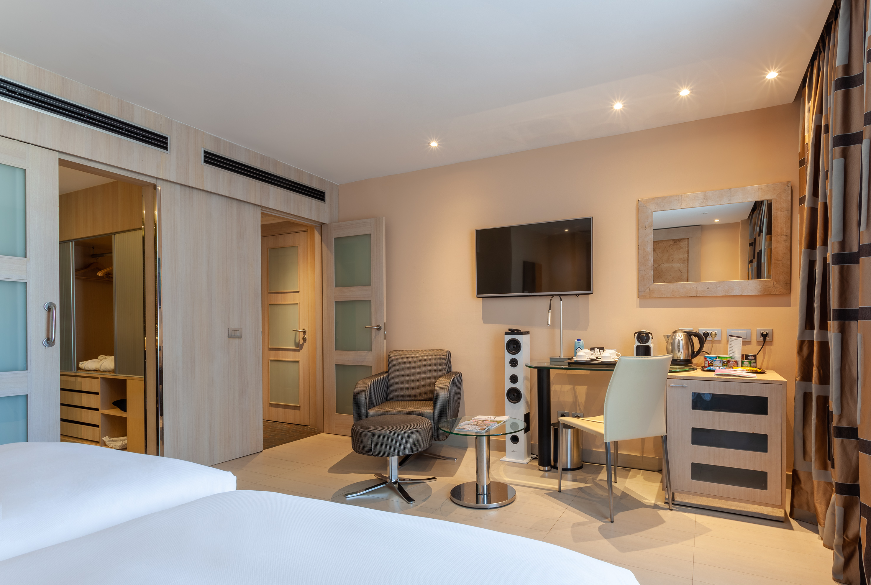 Modern hotel room with twin beds, desk, wall TV, armchair, and sliding closet.