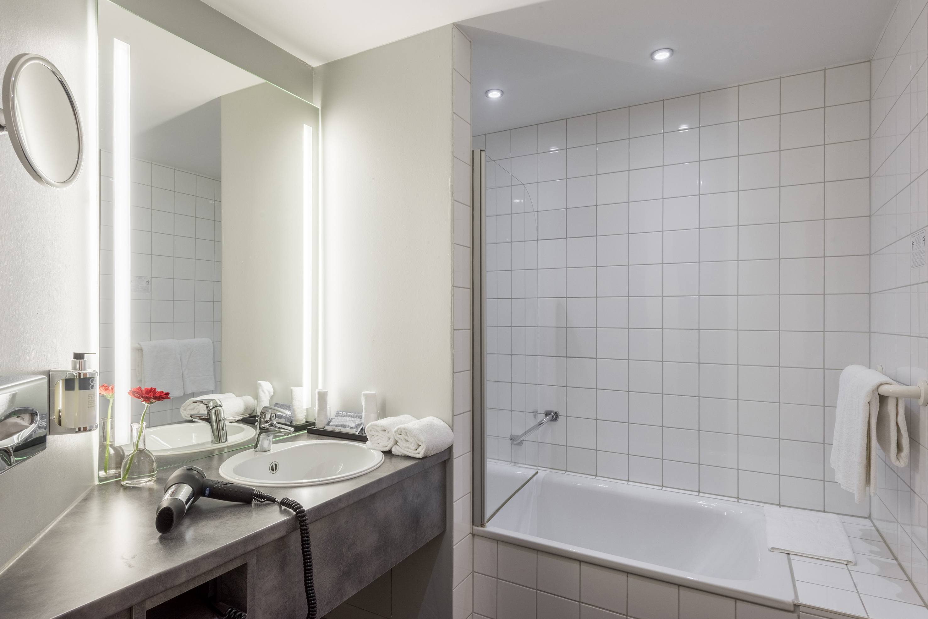 Modern hotel bathroom with sink, lighted mirror, hairdryer, bathtub, white tiles.