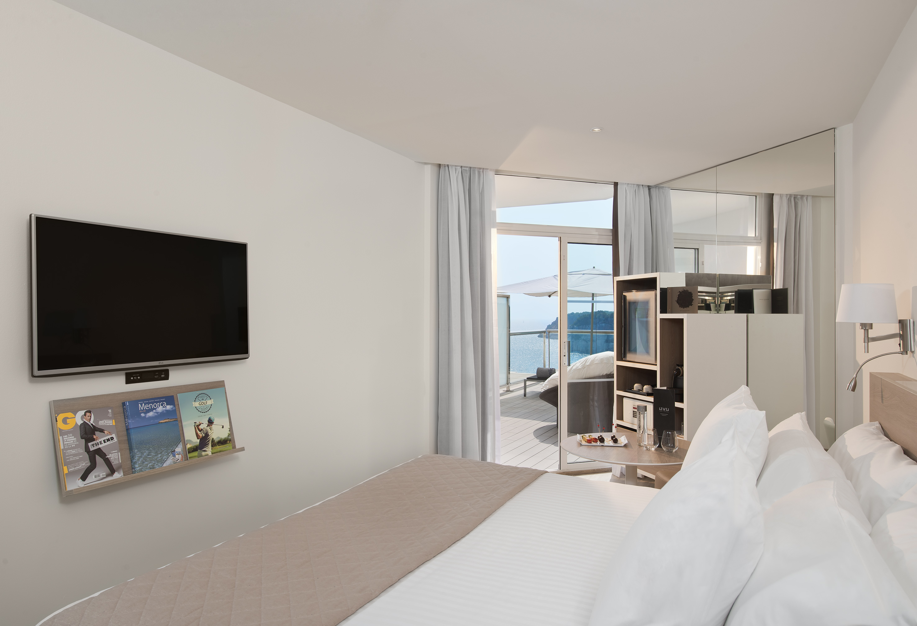Bright hotel room with bed, wall-mounted TV, and balcony overlooking the sea.