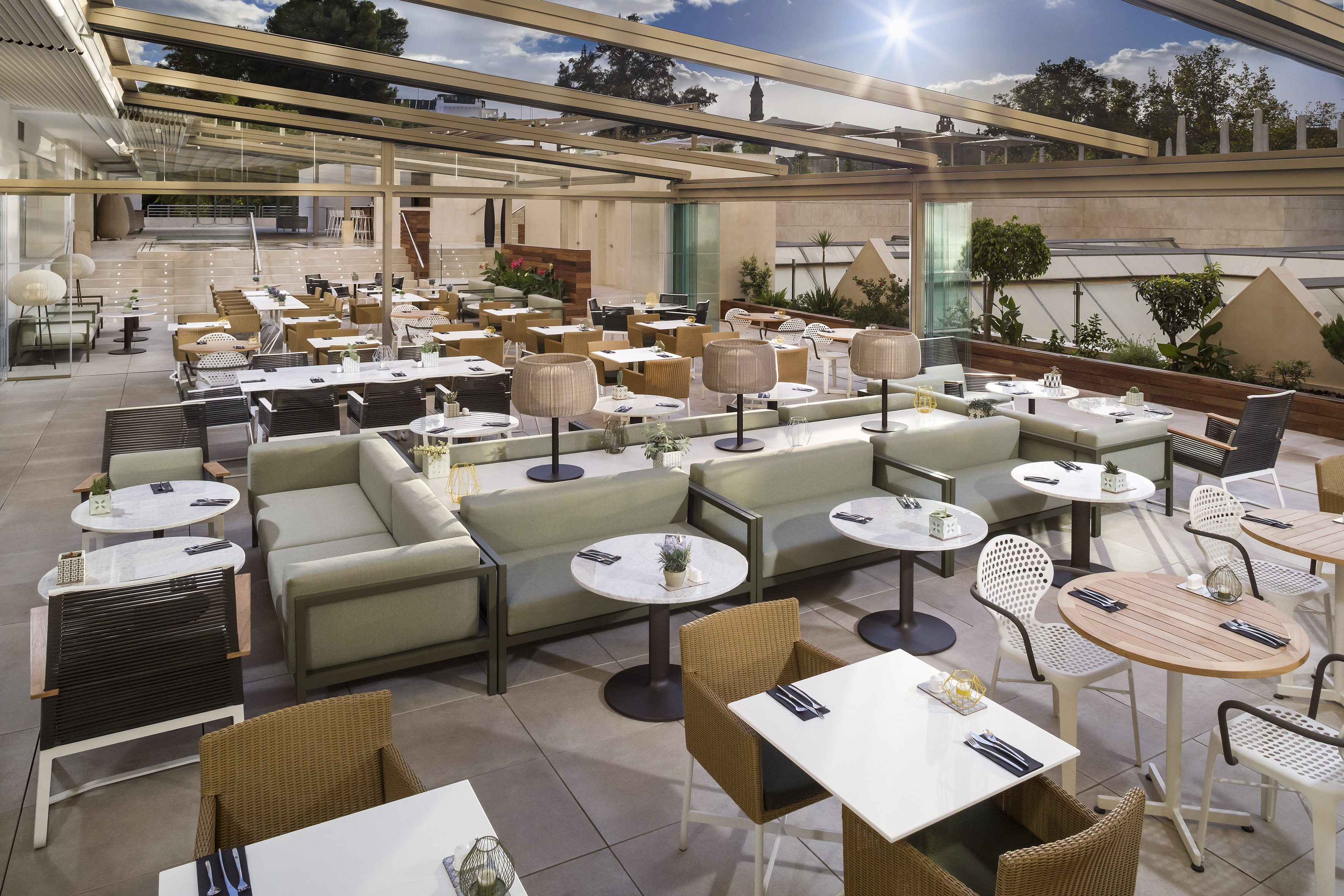 Modern outdoor restaurant patio with tables, sofas, and a sunny glass roof.