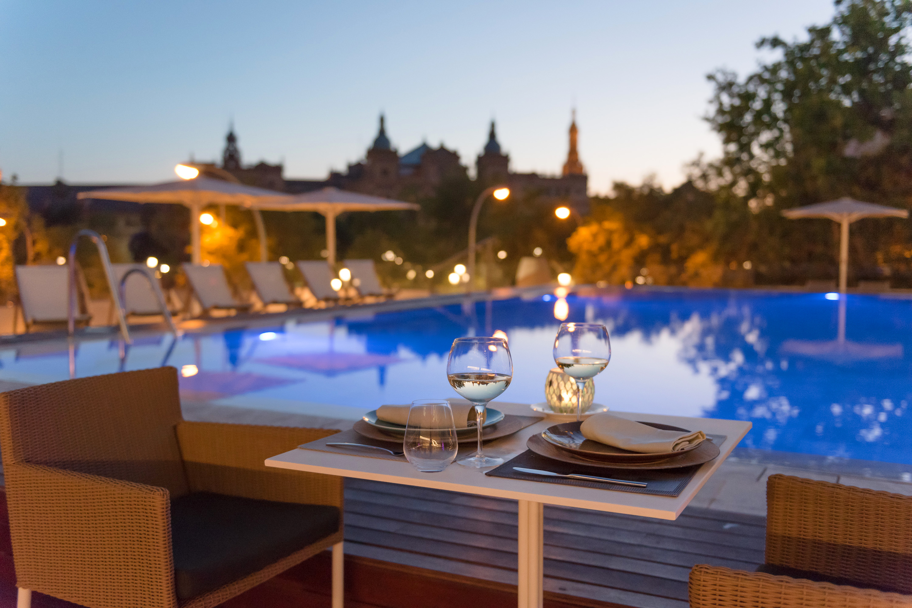 Poolside evening dining setup with wine, illuminated pool, city view.