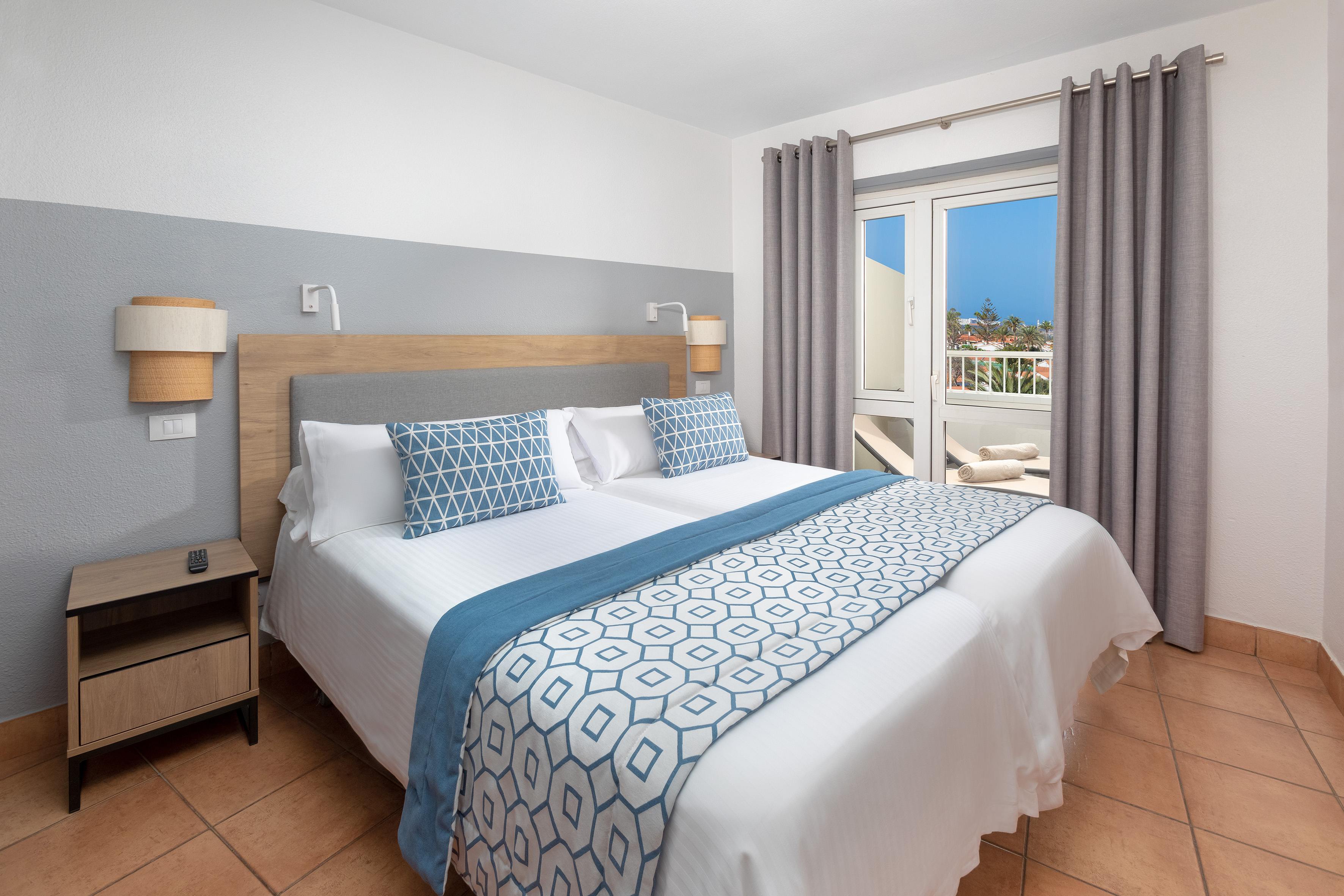 Modern resort bedroom with double bed, blue accents, balcony, and tile floor.
