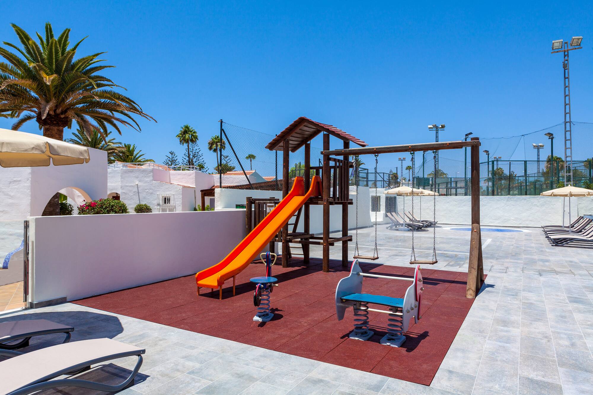 Sunny resort playground with orange slide, swings, and palm trees.