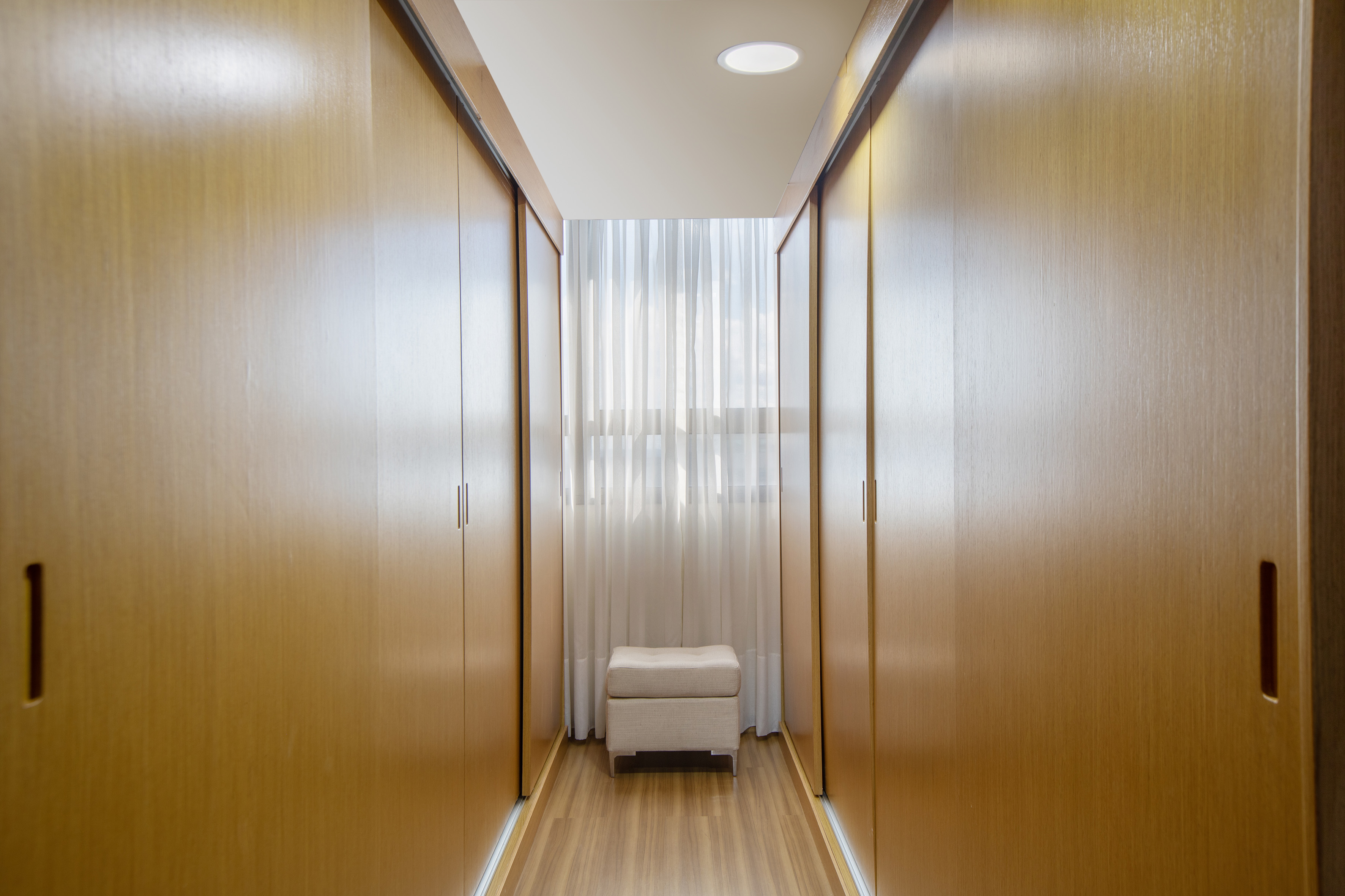 Modern walk-in closet with light wood wardrobes, ottoman, and sheer curtains.