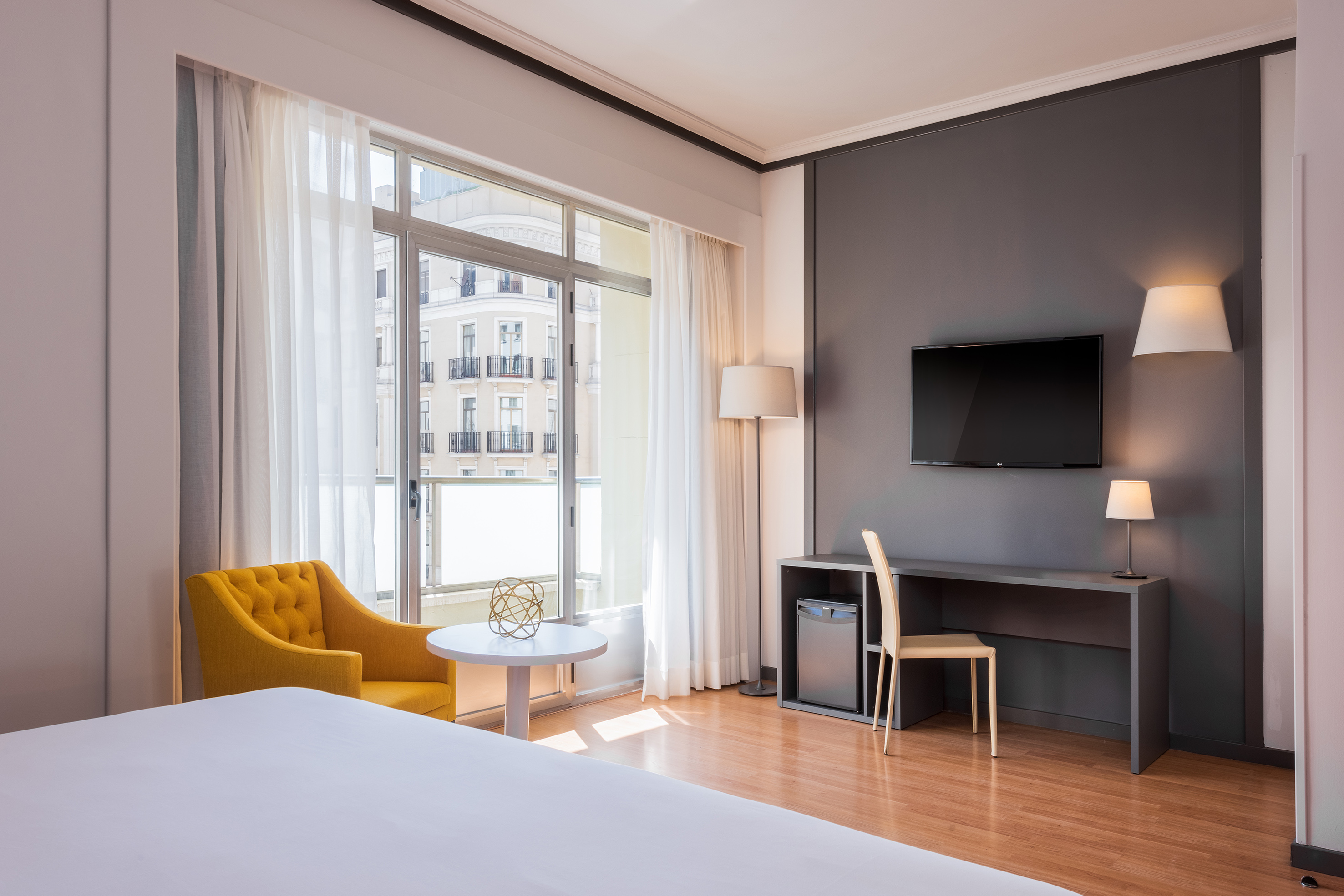 Modern hotel bedroom with yellow chair, city view window, desk, and TV.