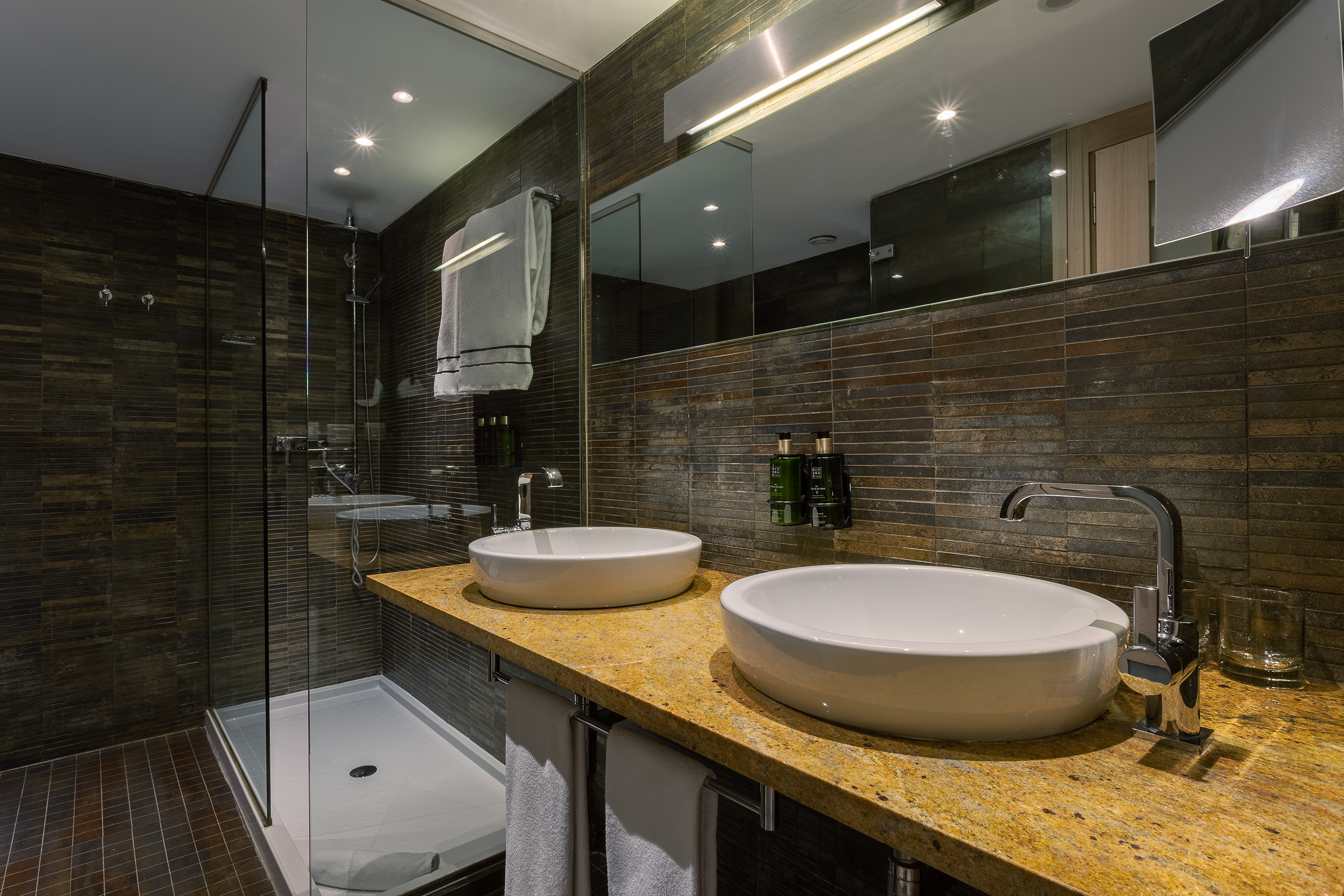 Modern bathroom: double vessel sinks, granite vanity, dark tiles, glass shower.