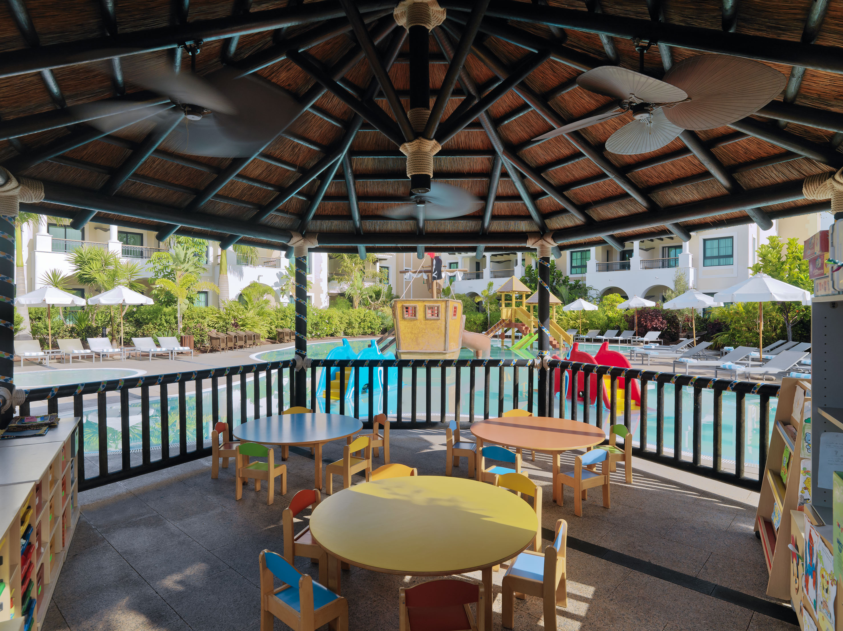 Resort kids' club: colorful tables, thatched gazebo, poolside, and water playground.