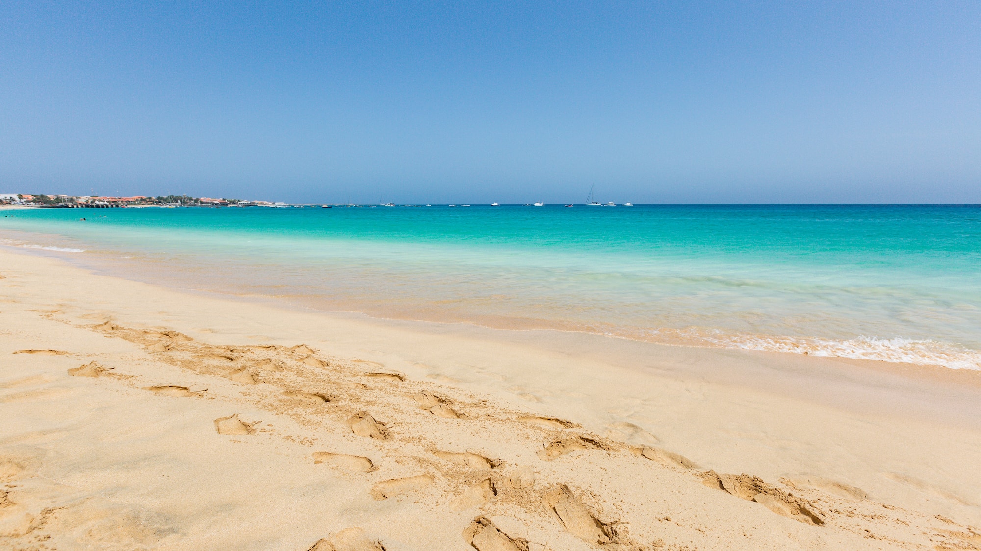 a sandy beach with blue water and boats in the background