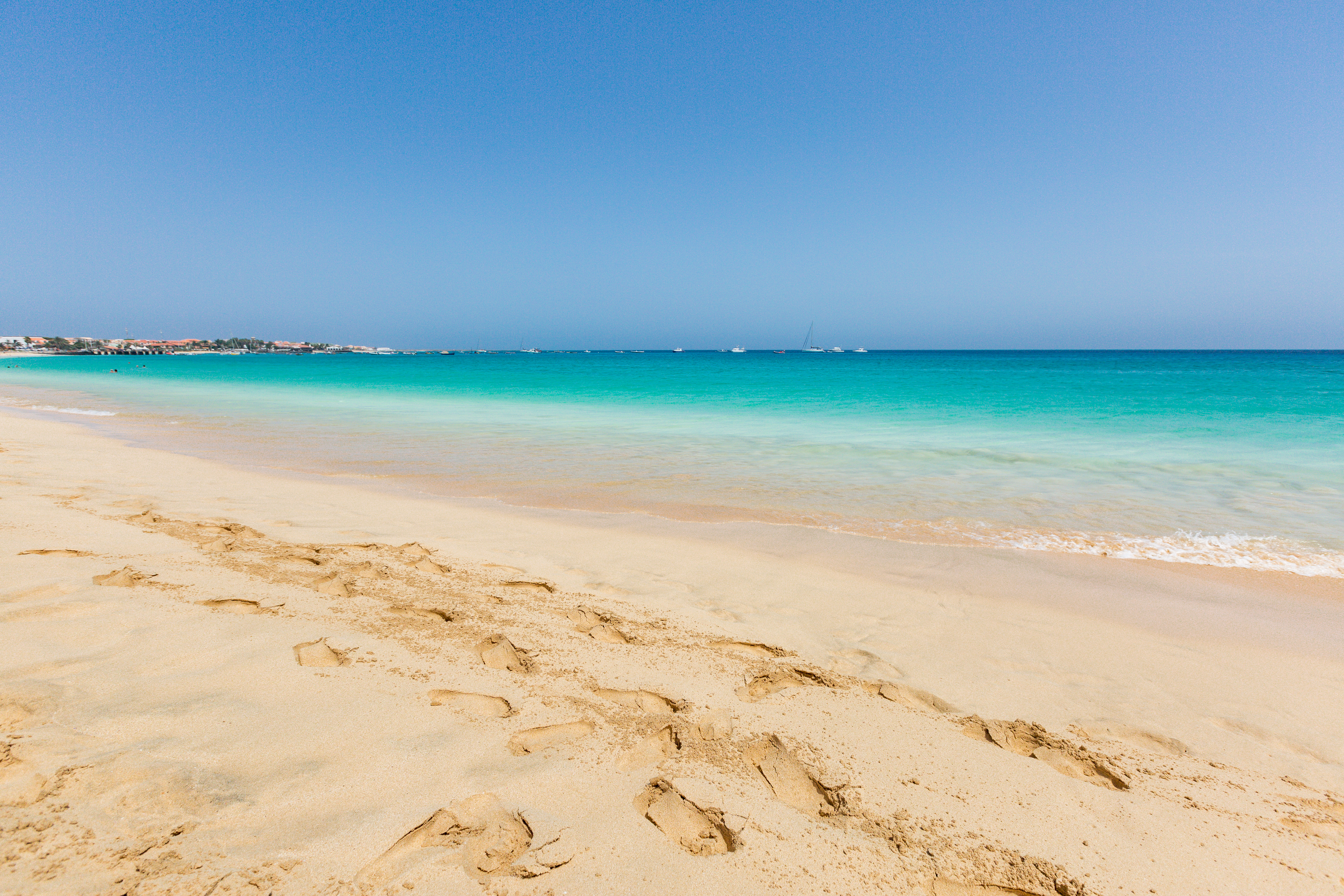 a sandy beach with blue water and boats in the background