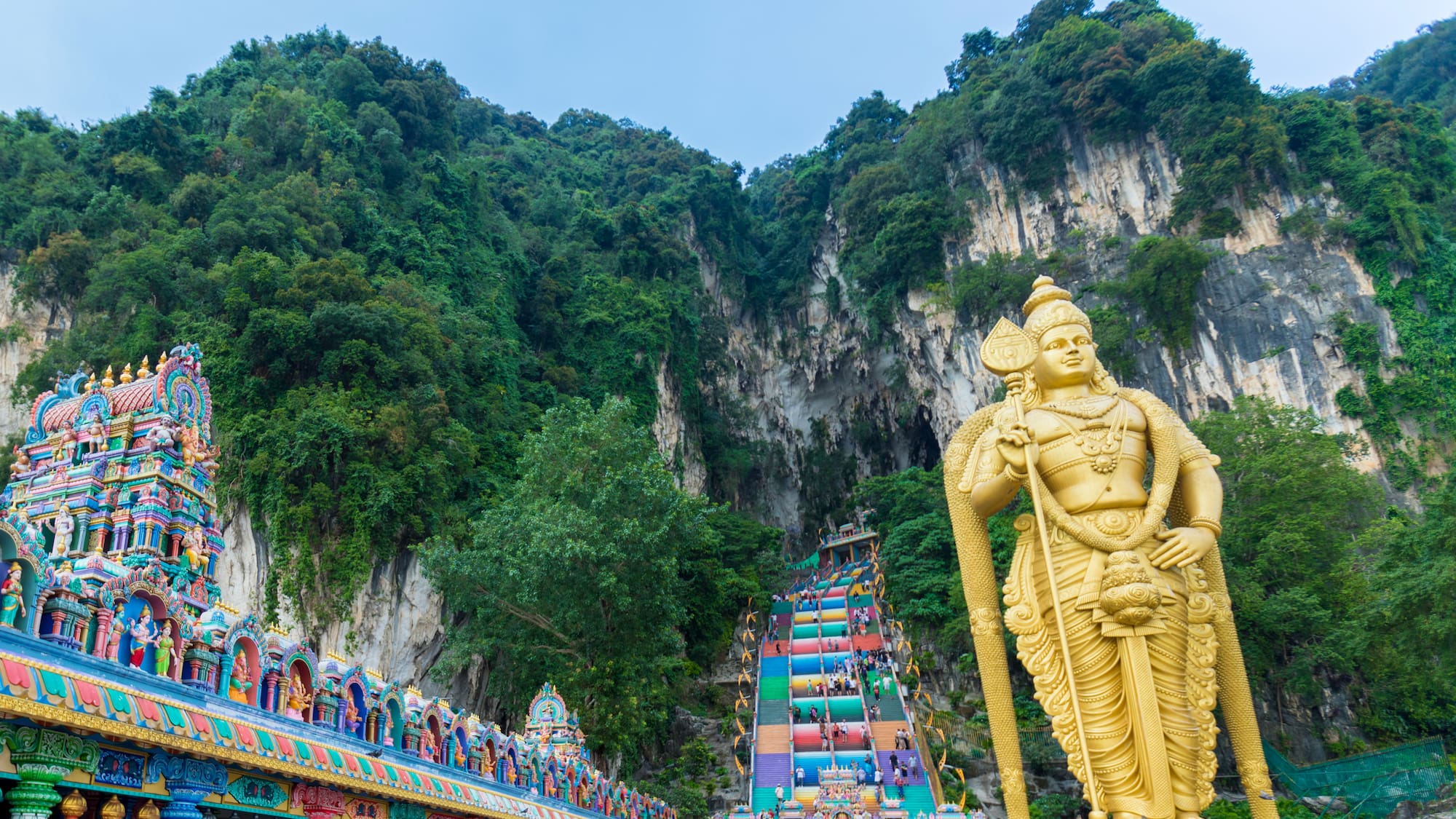 a statue of a man with a cane and a colorful staircase in front of Batu Caves