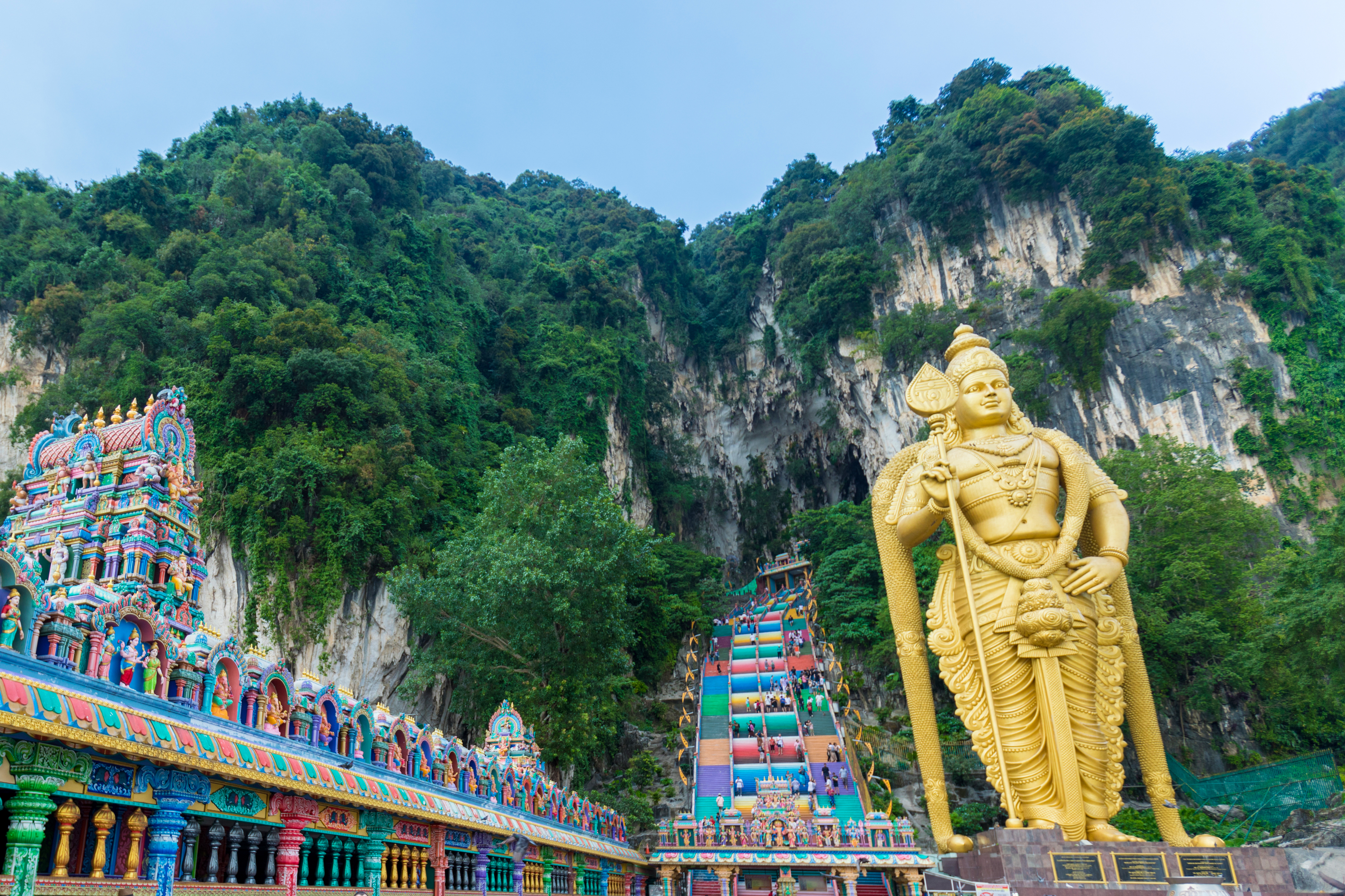 a statue of a man with a cane and a colorful staircase in front of Batu Caves