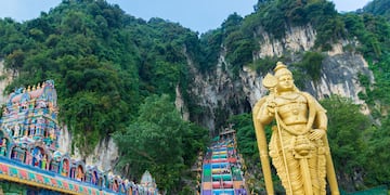 a statue of a man with a cane and a colorful staircase in front of Batu Caves