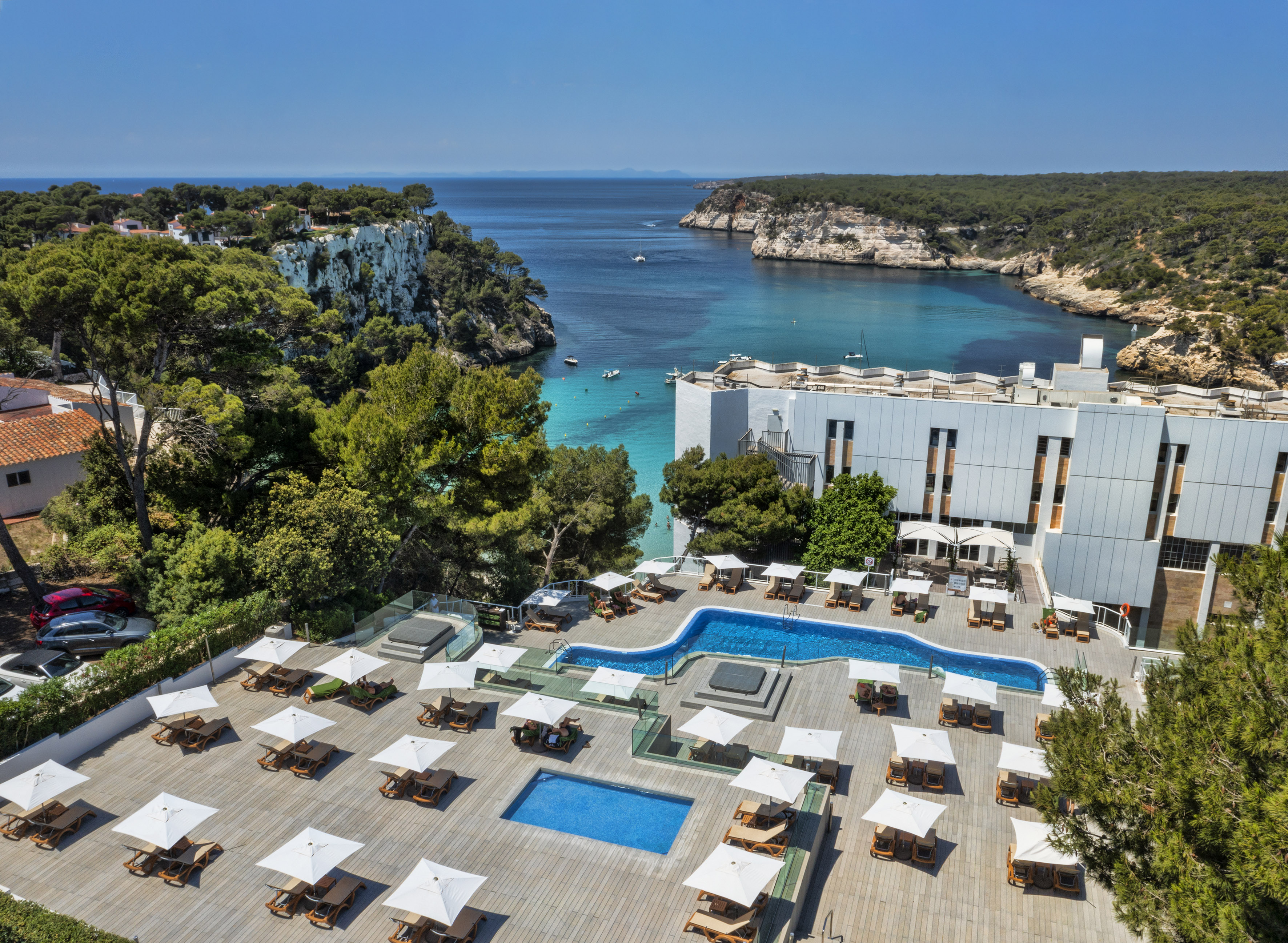 Modern resort, pools, sun loungers, overlooking clear blue Cala Galdana bay.