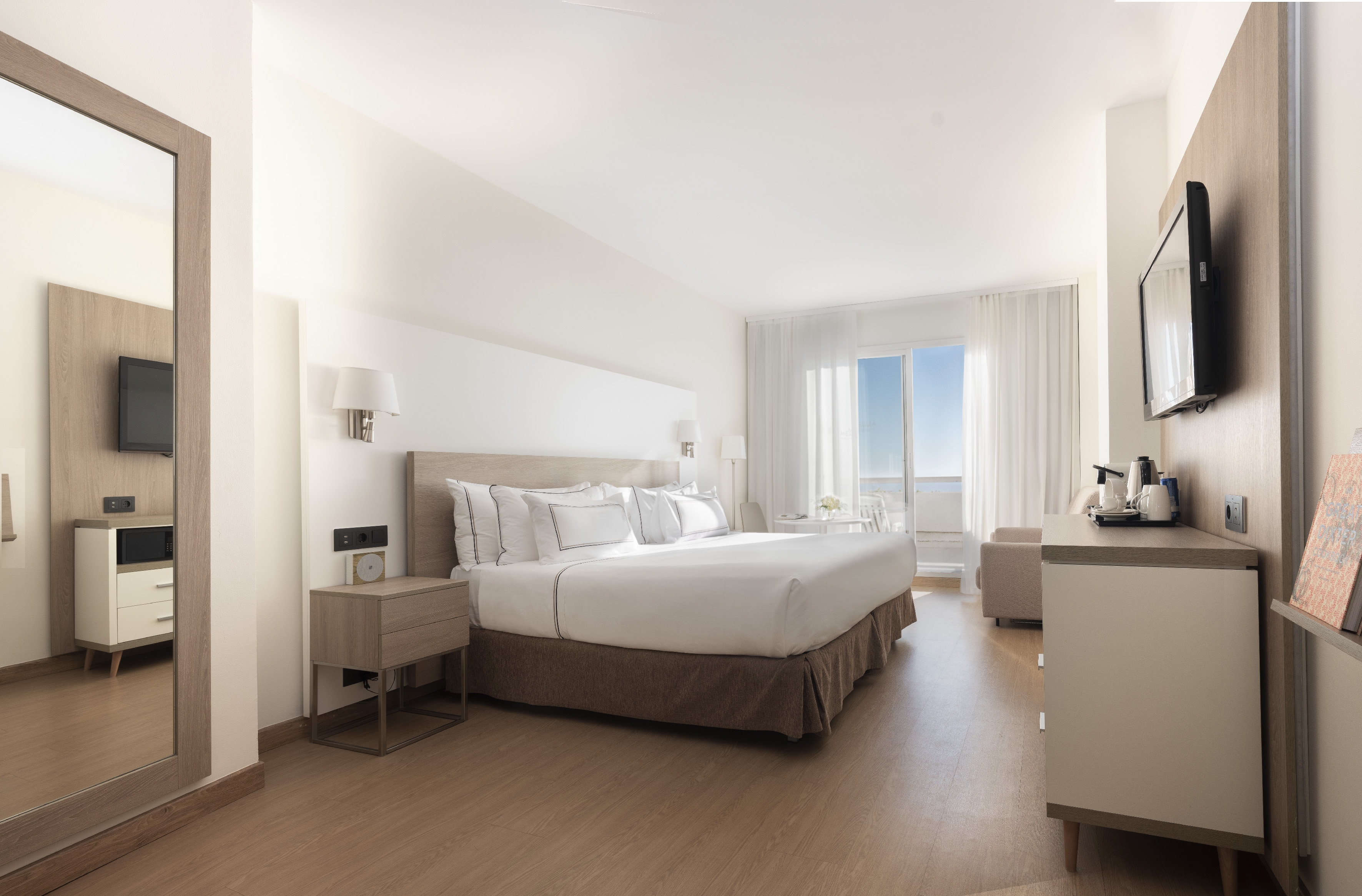 Bright hotel bedroom with king bed, balcony, and modern wooden furniture.