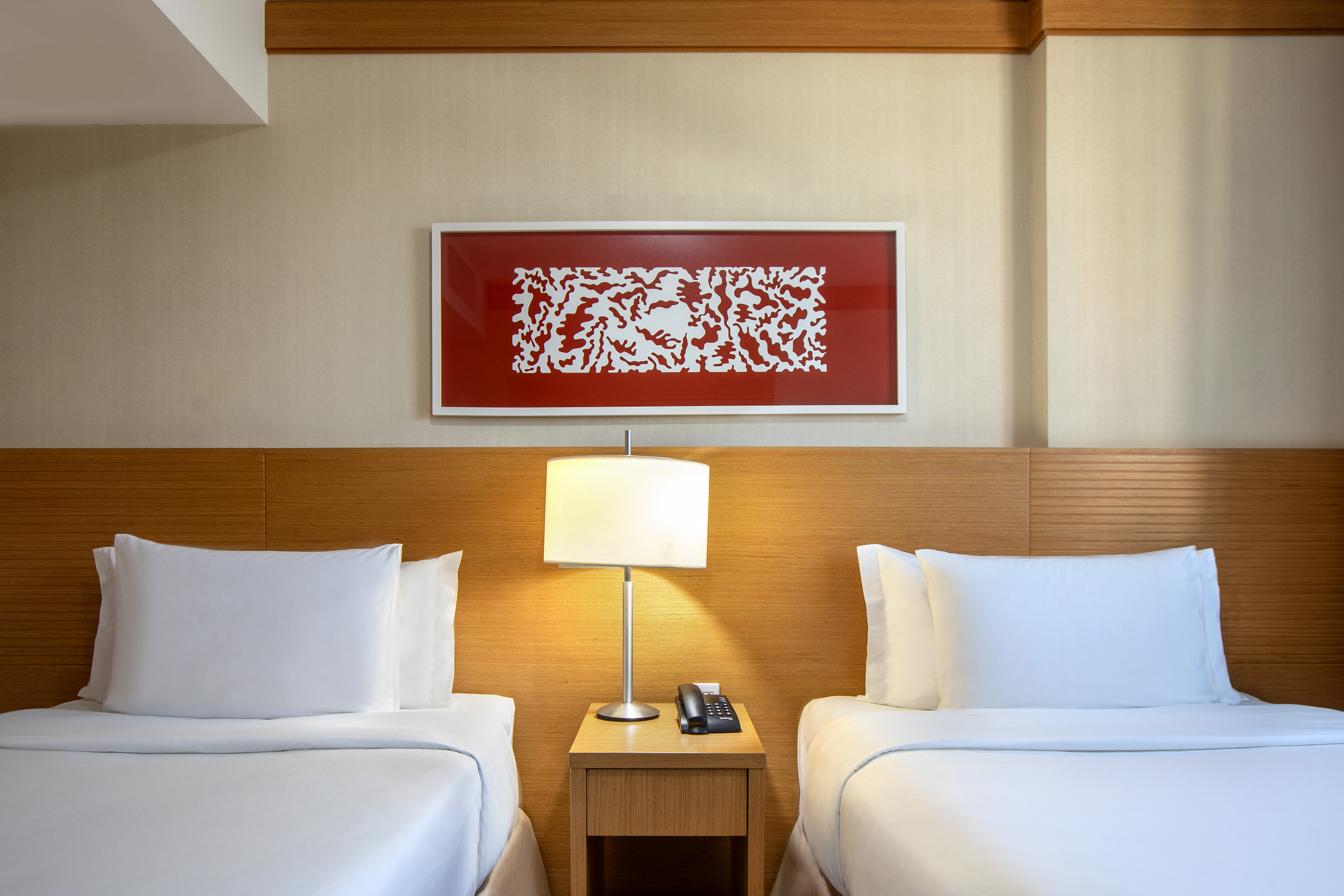 Bright hotel room with two twin beds, nightstand, lamp, and red abstract art.