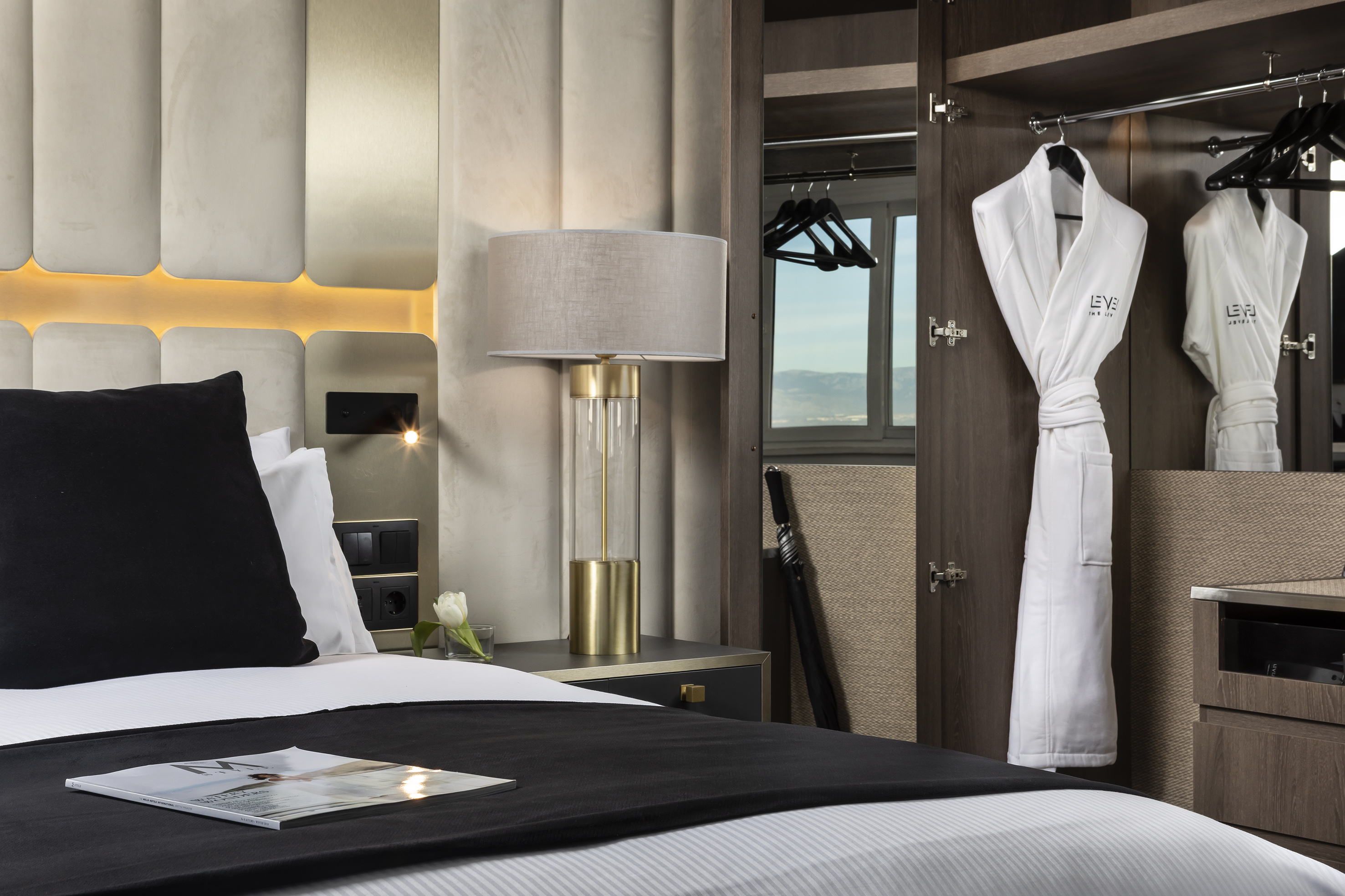 Modern hotel bedroom with lamp, bed, and open wardrobe with white bathrobes.