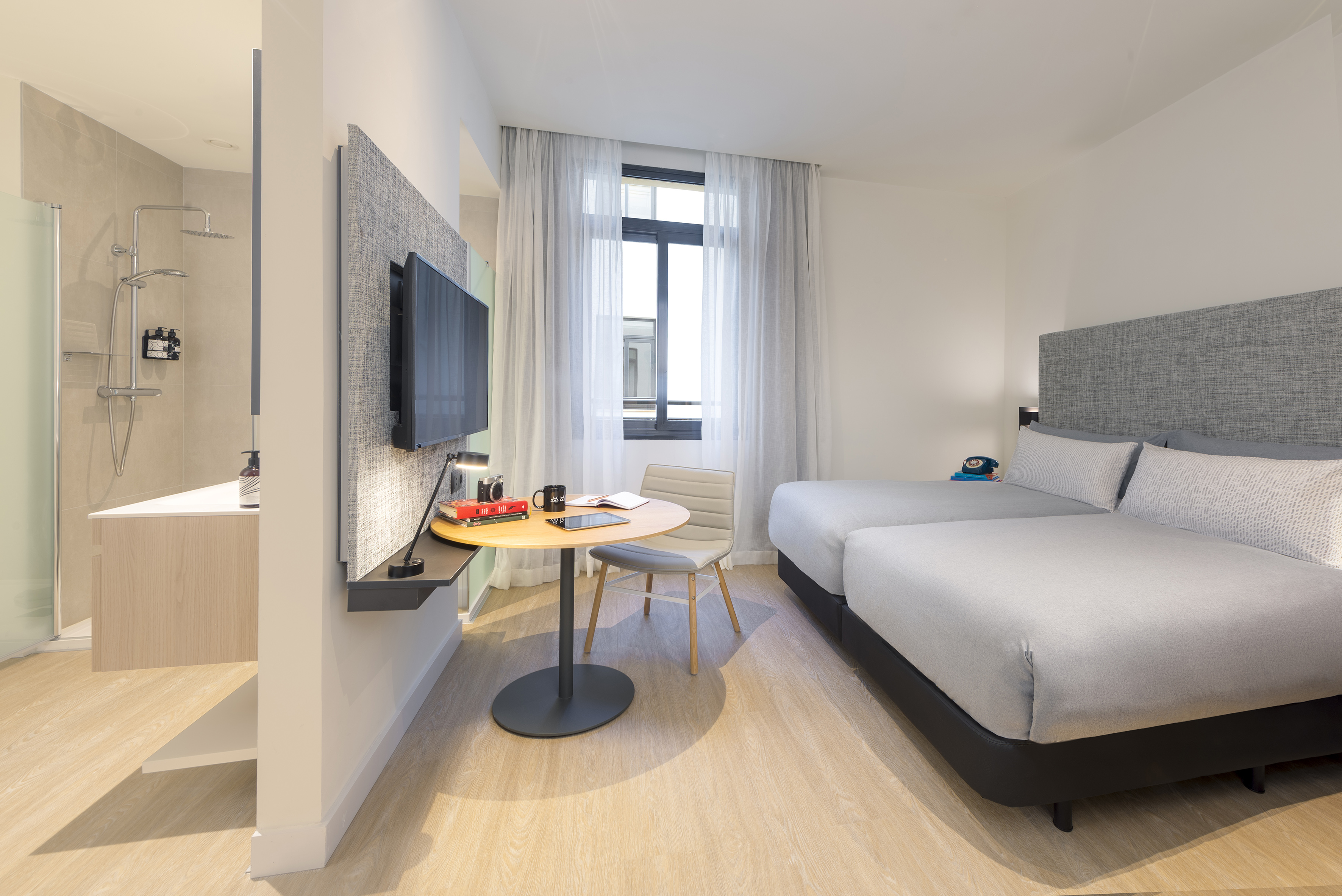 Modern hotel room with two twin beds, TV, work table, and open shower area.