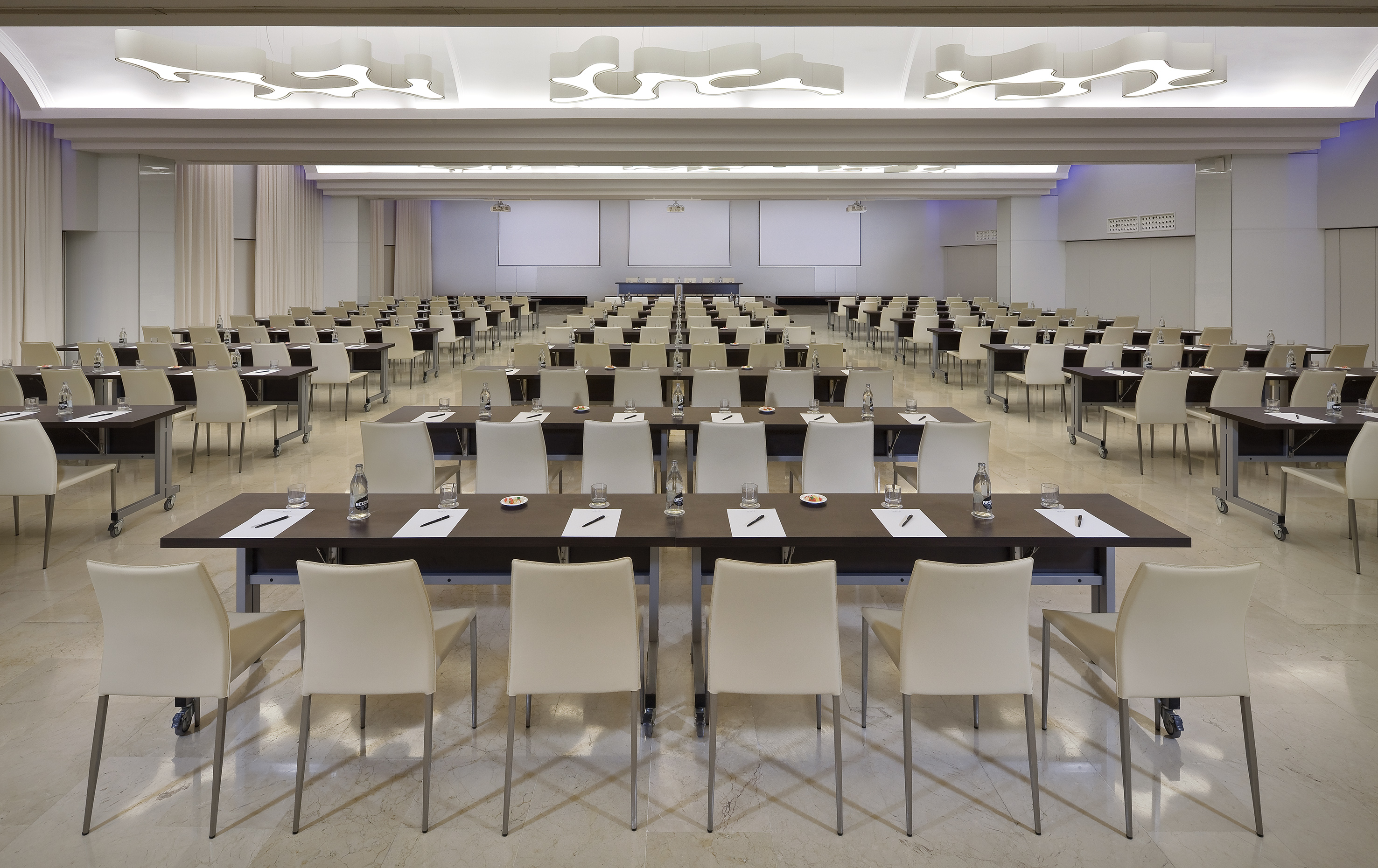 Spacious modern conference room with rows of tables, chairs, and projector screens.