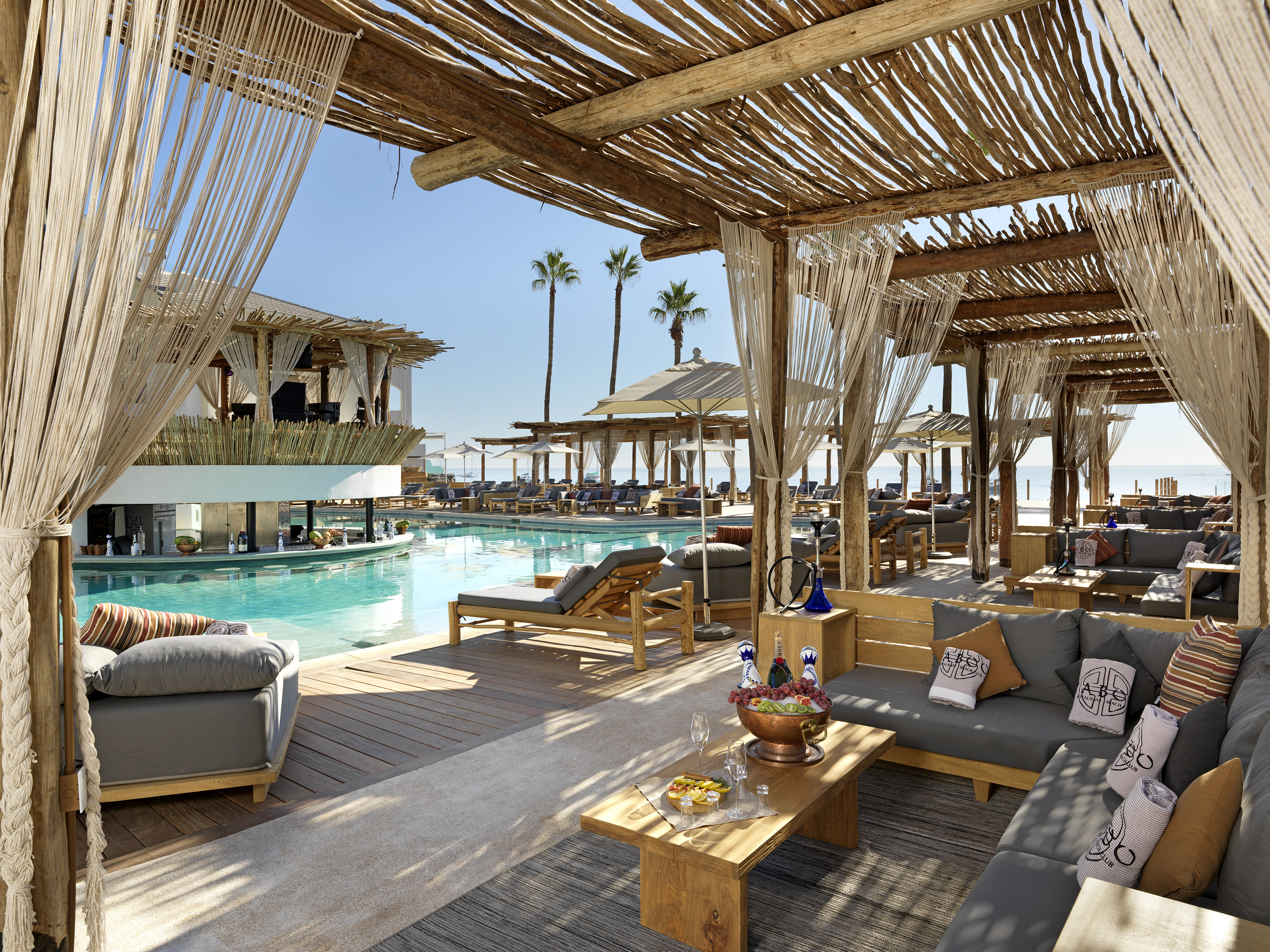 Luxury beach club lounge with pool, ocean view, palm trees, and shaded cabanas.