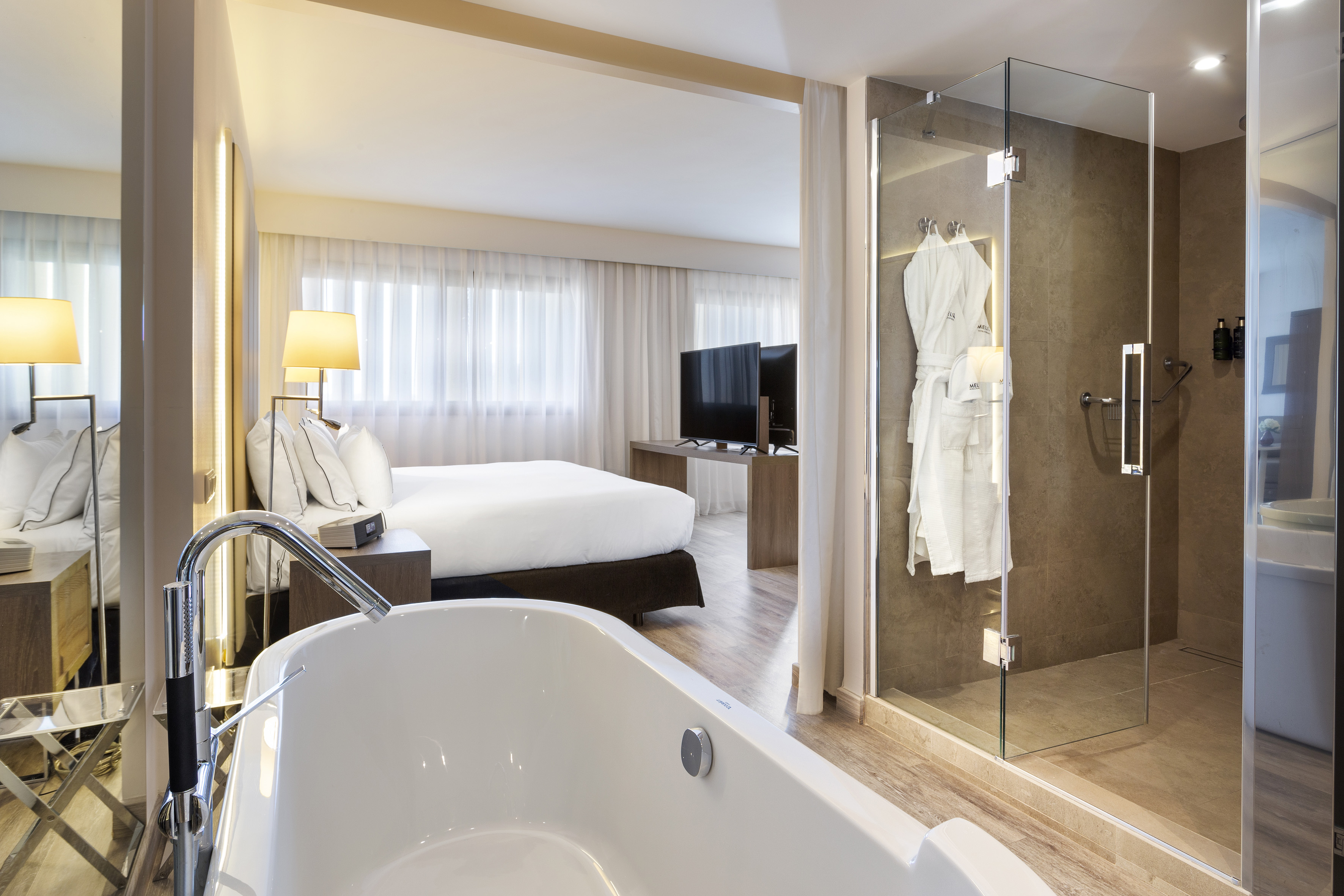 Luxurious hotel suite with deep soaking tub, bed, and glass walk-in shower.