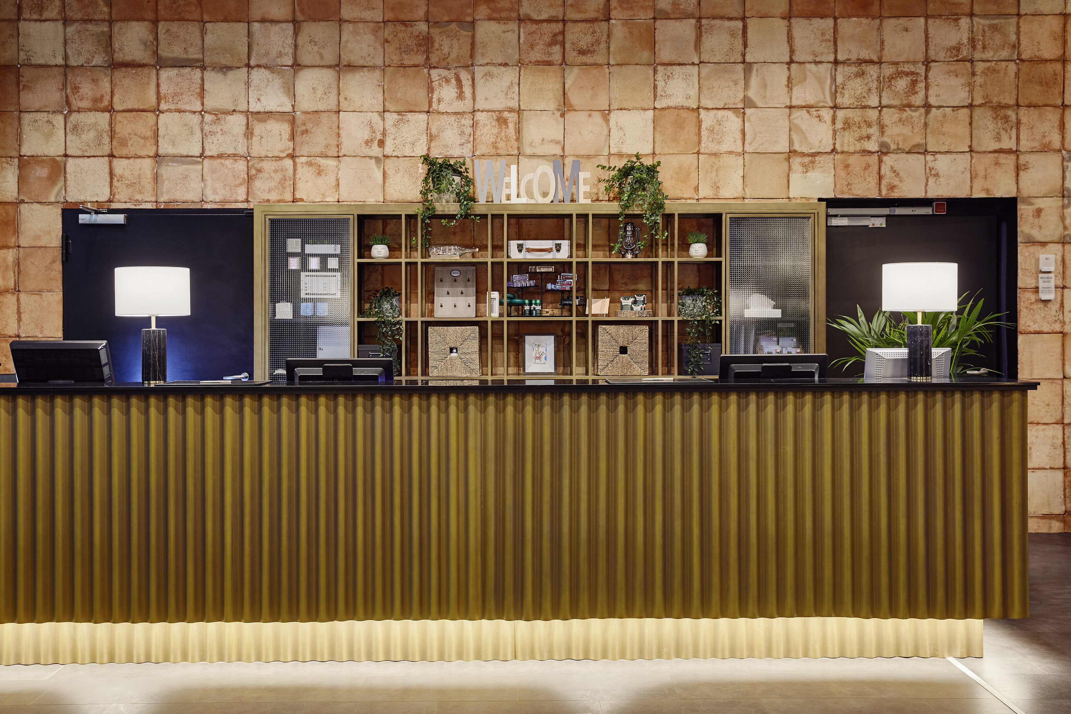 Hotel reception desk with gold corrugated front, welcome sign, and terracotta wall.