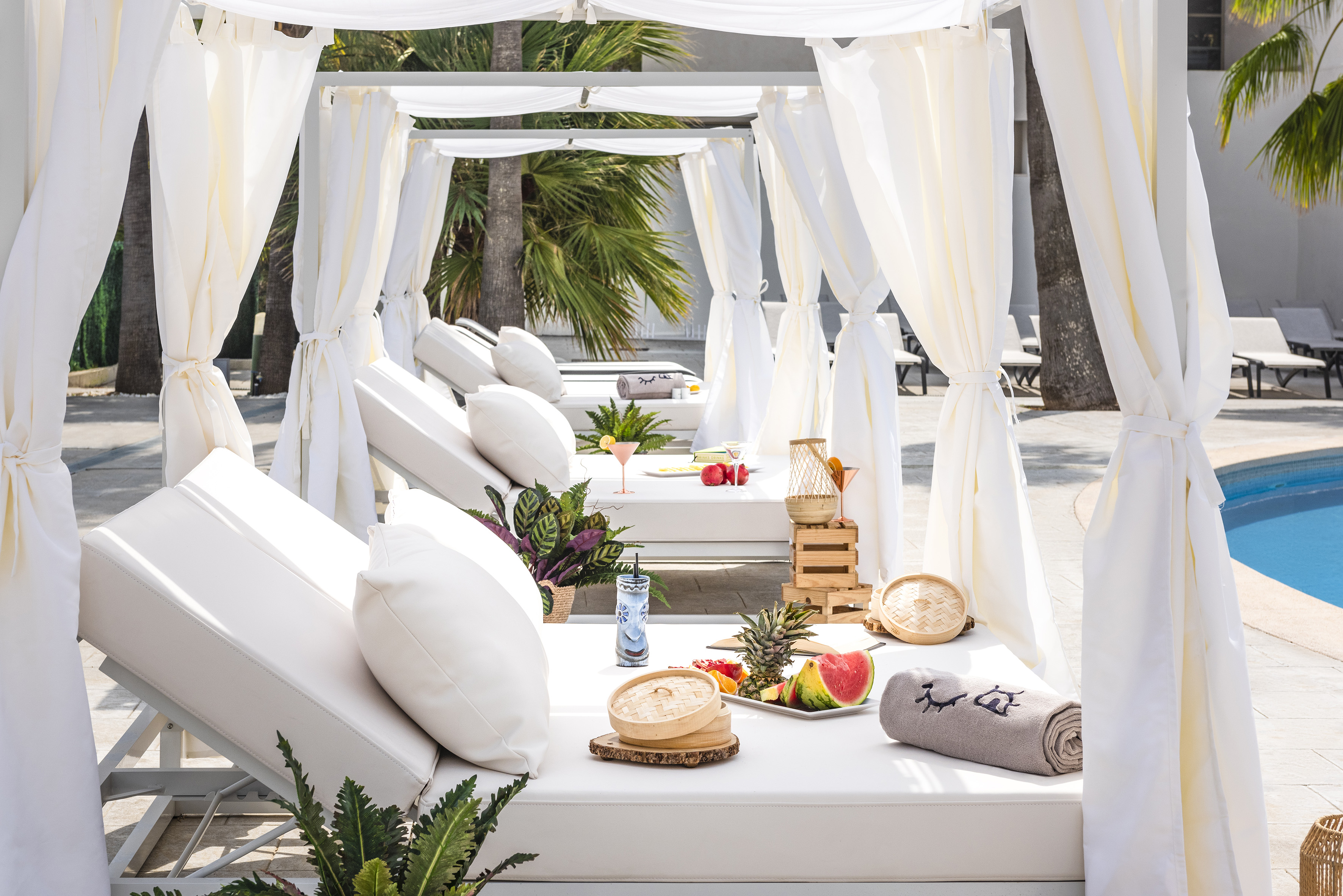 White cabana daybeds by resort pool with tropical fruit and drinks.